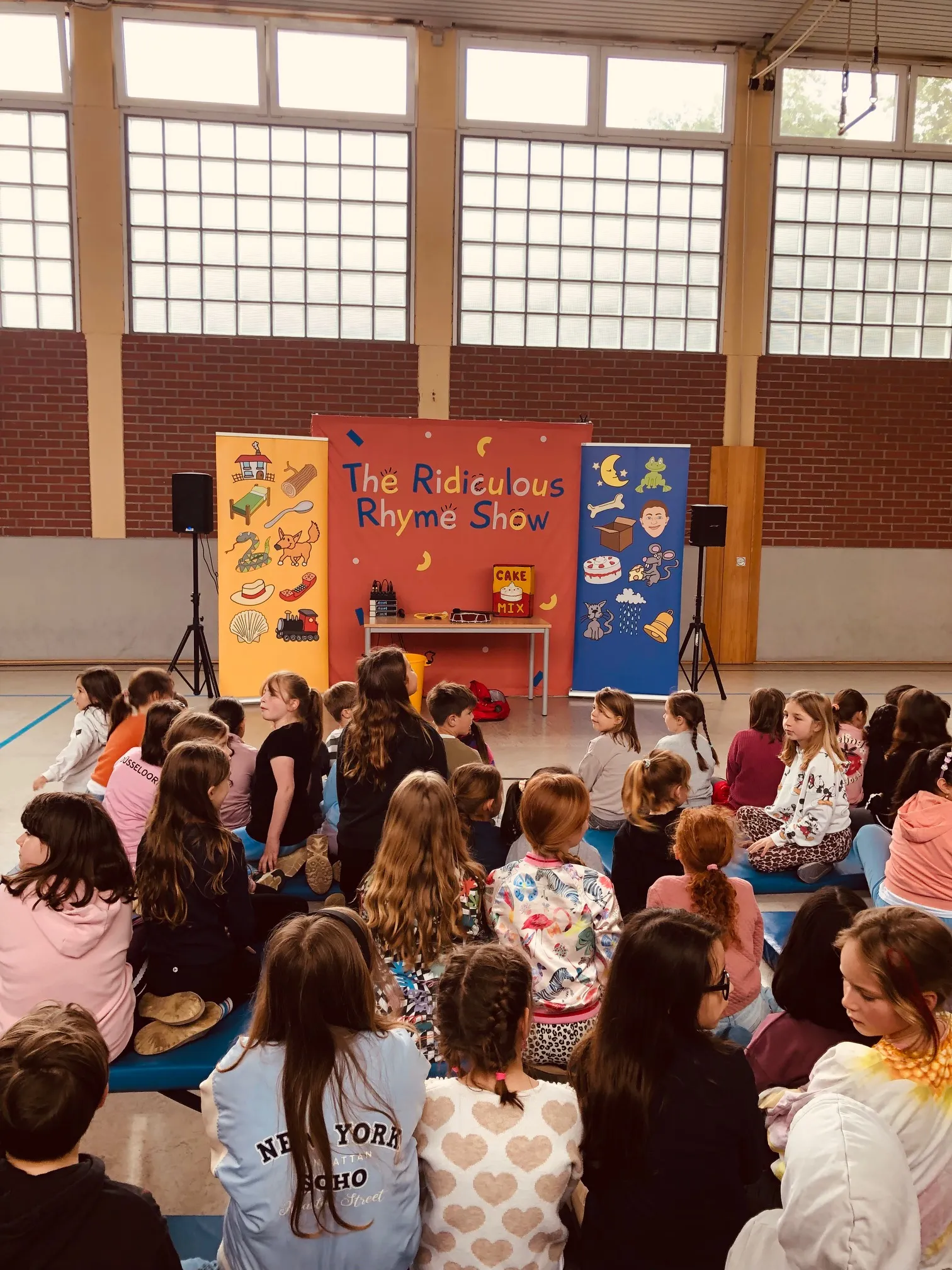 Viele Kinder sitzen in einer Turnhalle vor einer Bühne mit bunten Bannern und Schriftzug "The Ridiculous Rhyme Show".