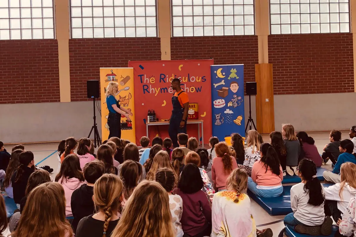 Kinder sitzen in einer Turnhalle vor zwei Darstellern und bunten Bühnenbannern.