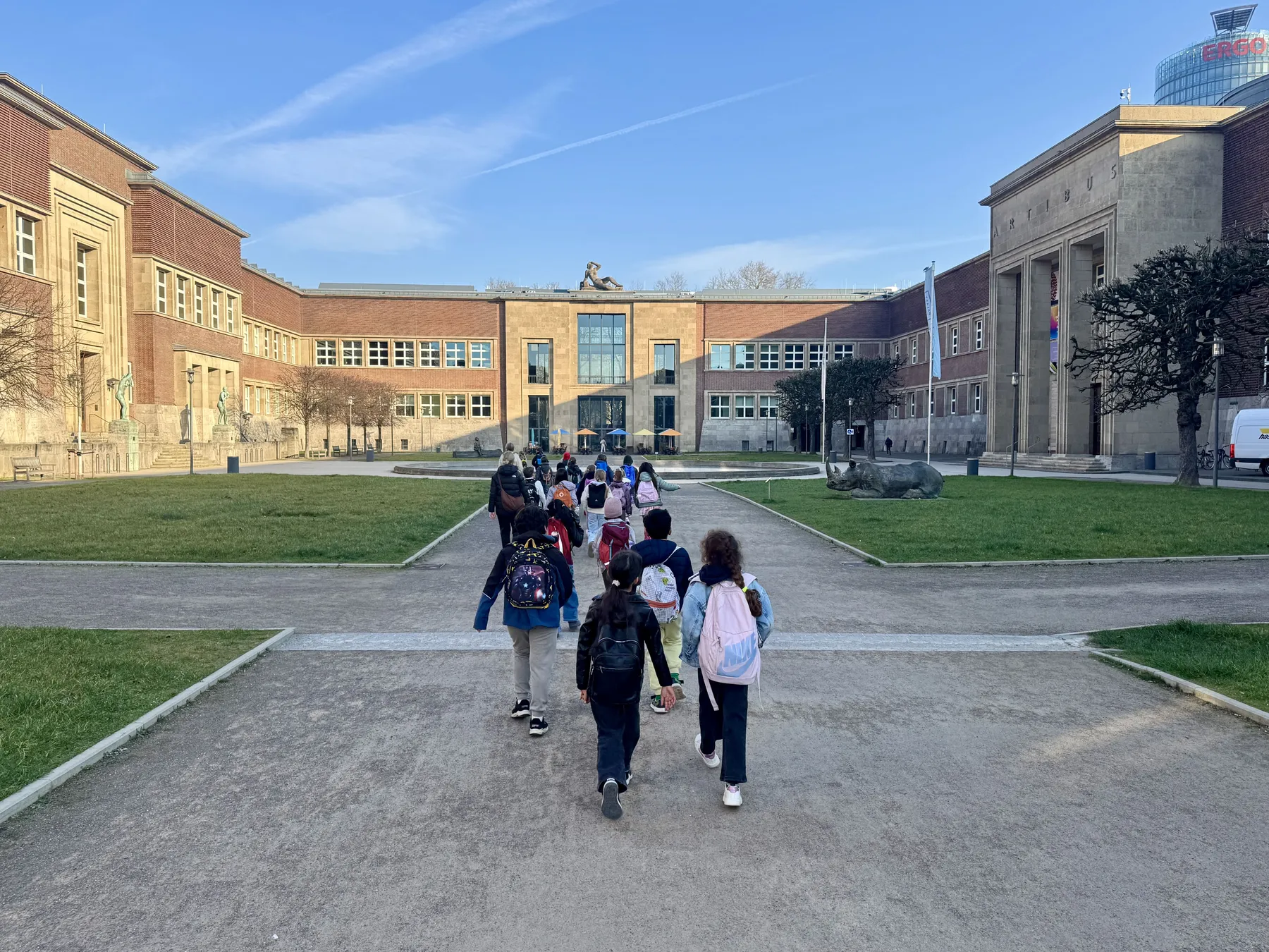 Schulkinder mit Rucksäcken gehen auf ein großes Museumsgebäude mit Statuen und Nashornskulptur zu