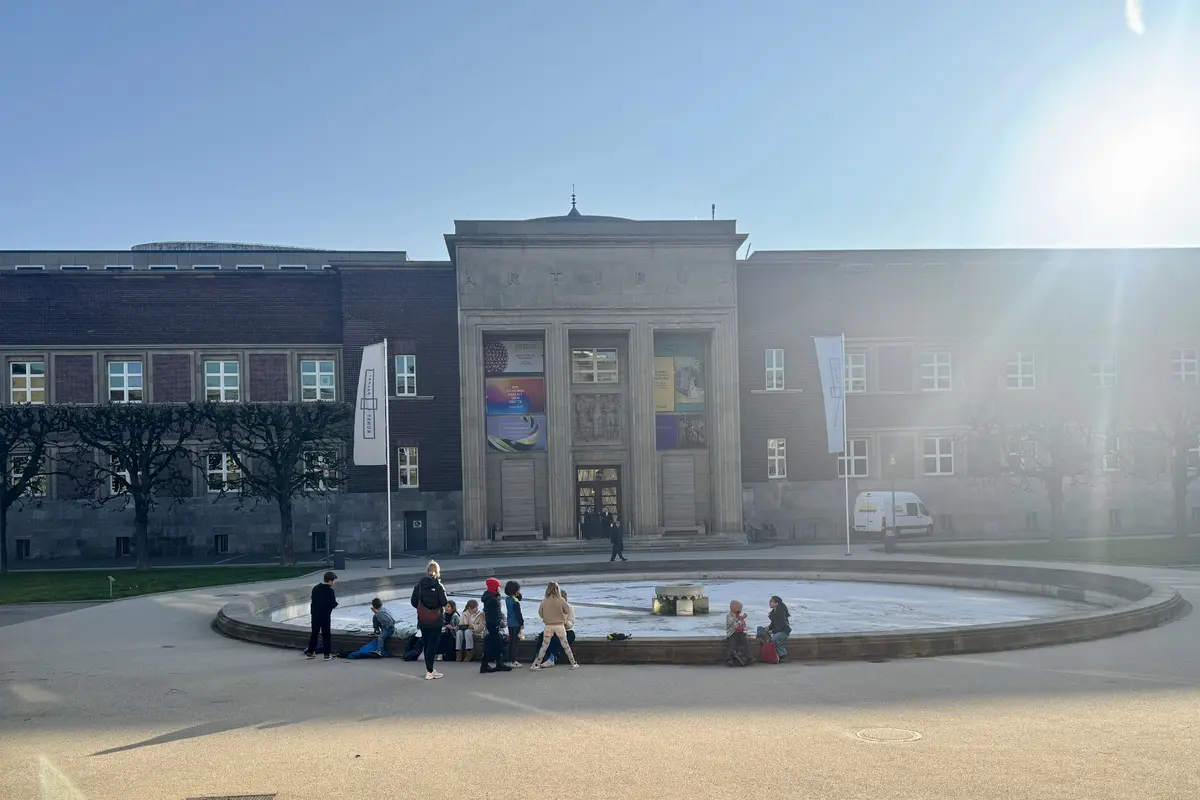 Großes Museumgebäude mit Säulenportal, davor runder Brunnen und eine Gruppe Kinder am Brunnenrand, Sonnenschein