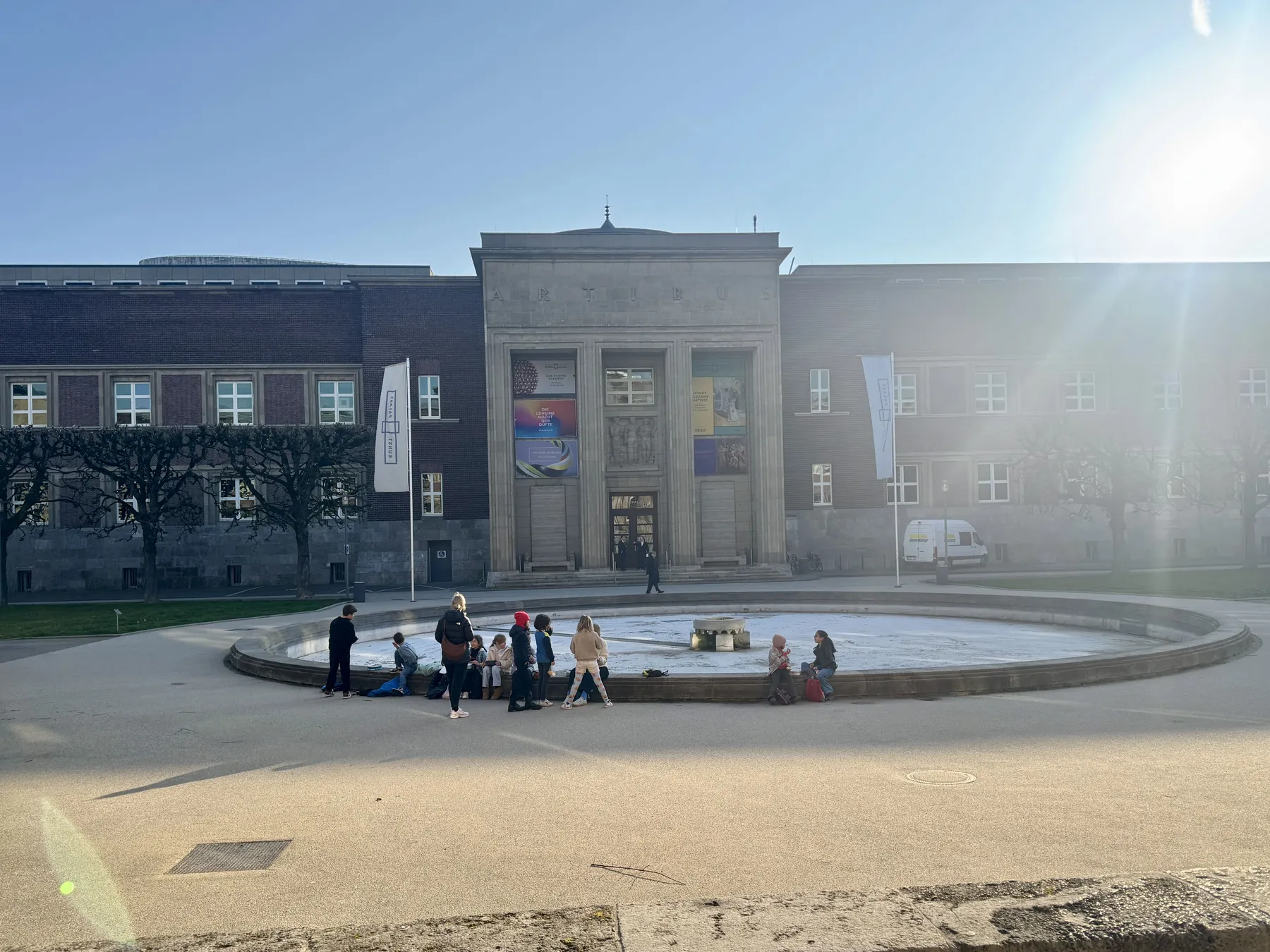 Großes Museumgebäude mit Säulenportal, davor runder Brunnen und eine Gruppe Kinder am Brunnenrand, Sonnenschein