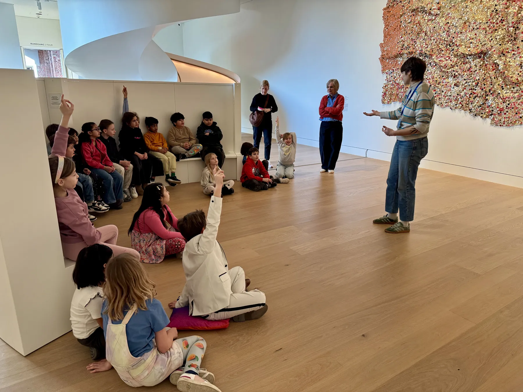 Kinder sitzen auf dem Boden vor einer Dozentin im Museum; rechts eine große, bunte, strukturierte Wandinstallation