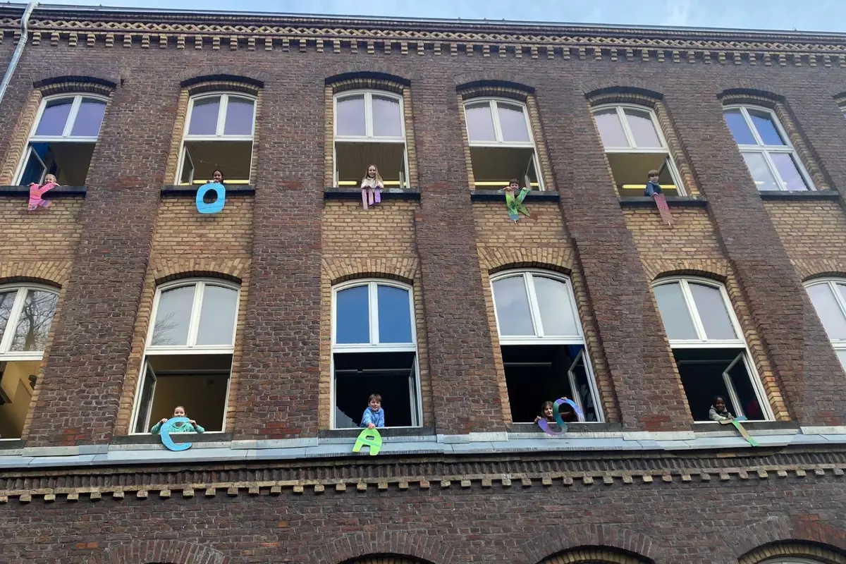 Backsteingebäude mit Kindern an geöffneten Fenstern, die bunte Papierbuchstaben aus den Fenstern halten