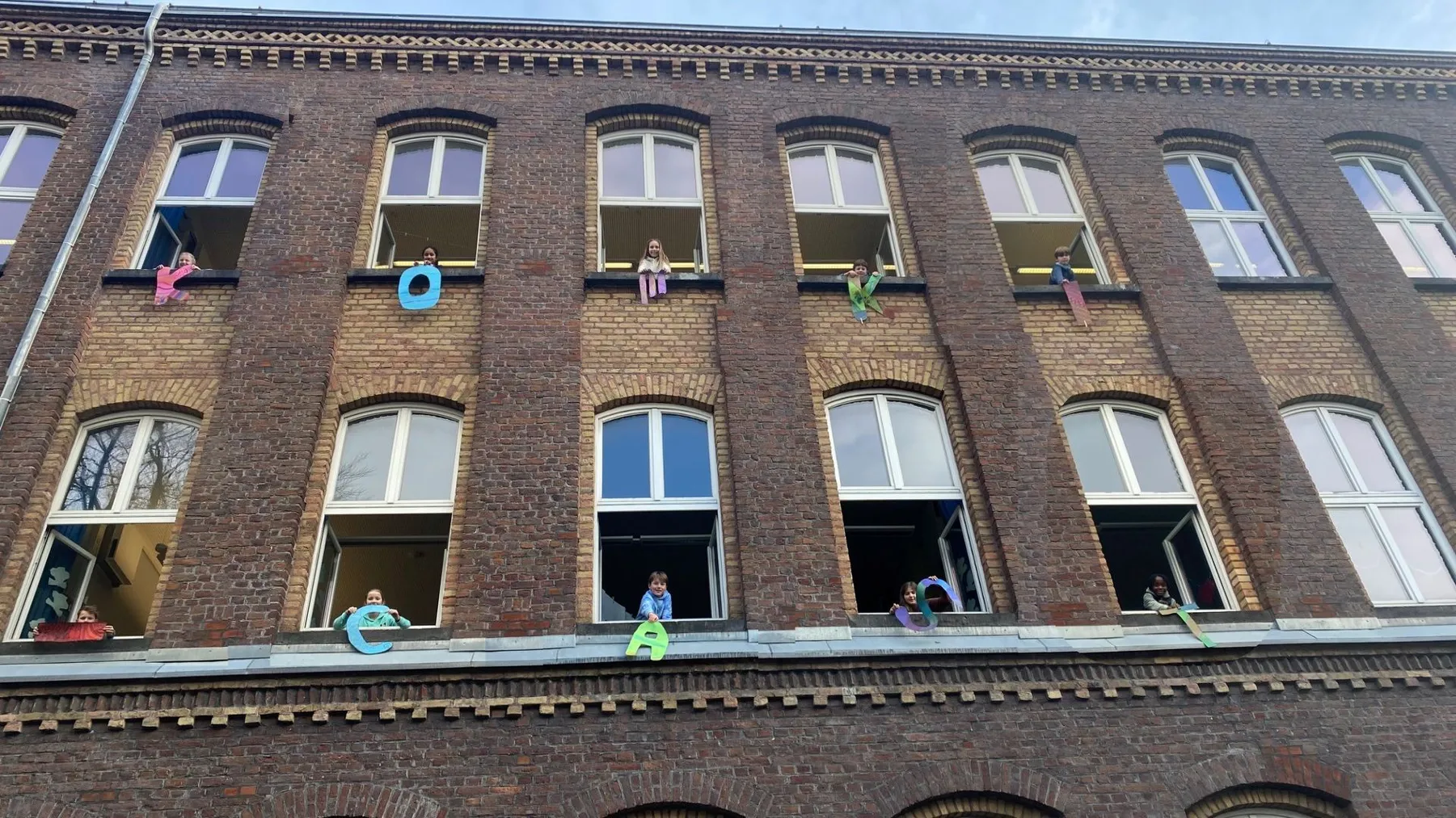 Backsteingebäude mit Kindern an geöffneten Fenstern, die bunte Papierbuchstaben aus den Fenstern halten