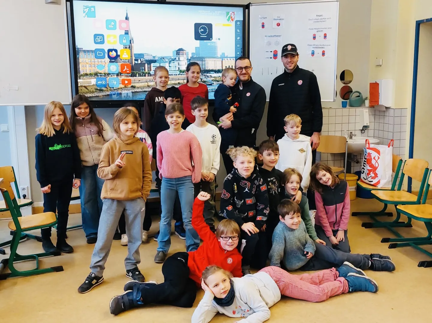 Etwa 16 Kinder und zwei Männer posieren in einem Klassenraum vor interaktivem Bildschirm.