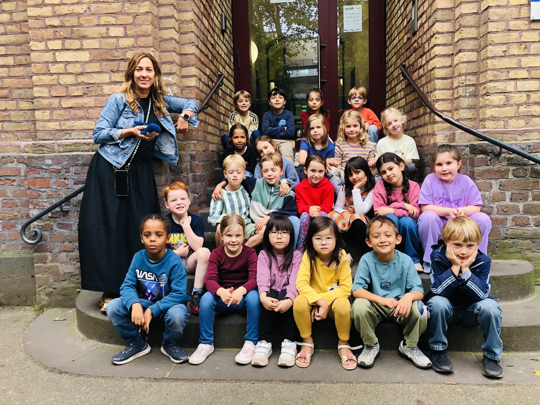 Lehrerin mit einer Gruppe Grundschulkinder auf Treppen vor einem ziegelroten Backsteingebäude