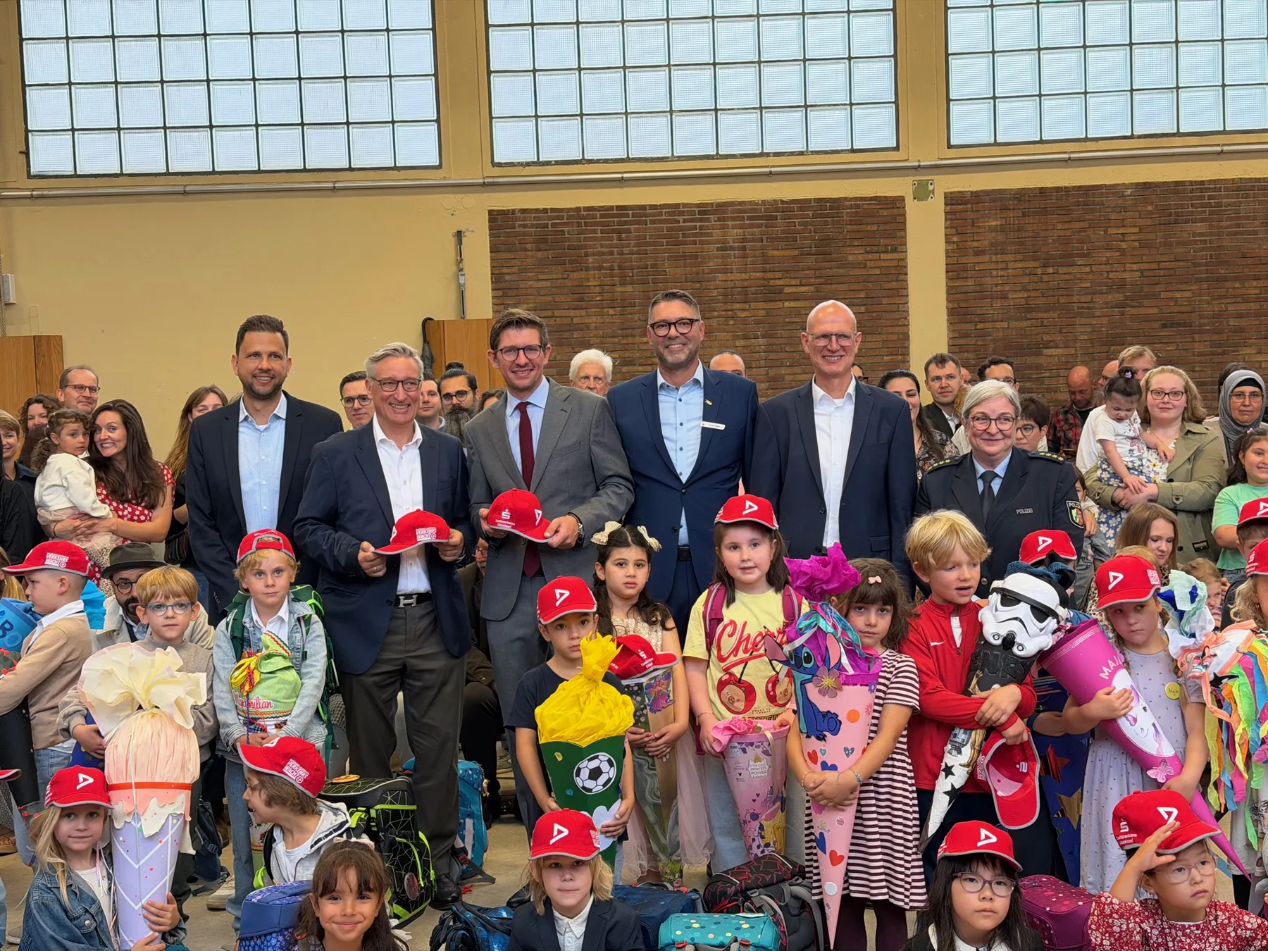 Gruppenfoto: Kinder mit bunten Schultüten und roten Kappen vor mehreren Erwachsenen in einer Halle