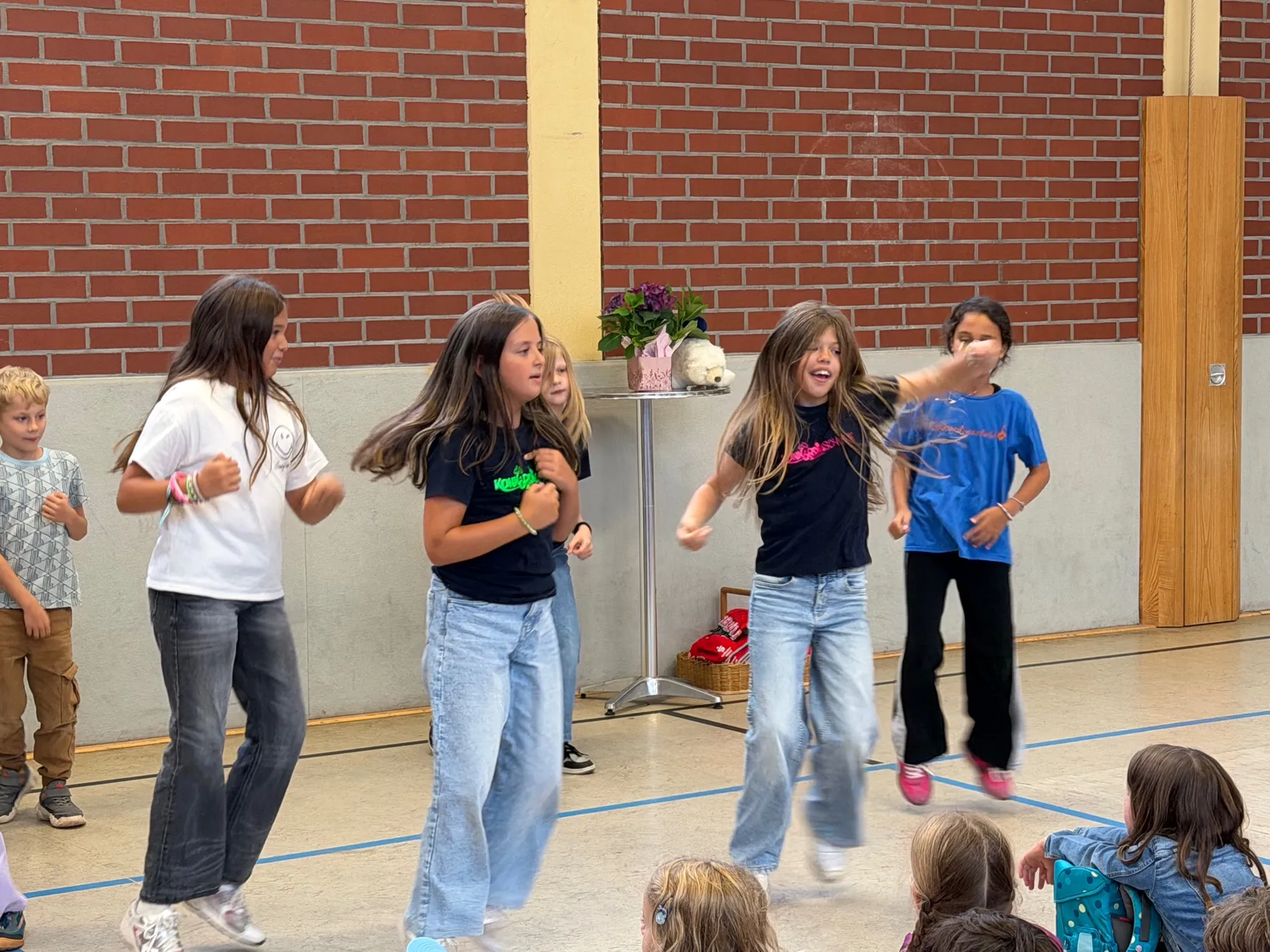 Fünf Kinder springen und tanzen in einer Turnhalle vor roter Ziegelwand, ein Tisch mit Blumen im Hintergrund.