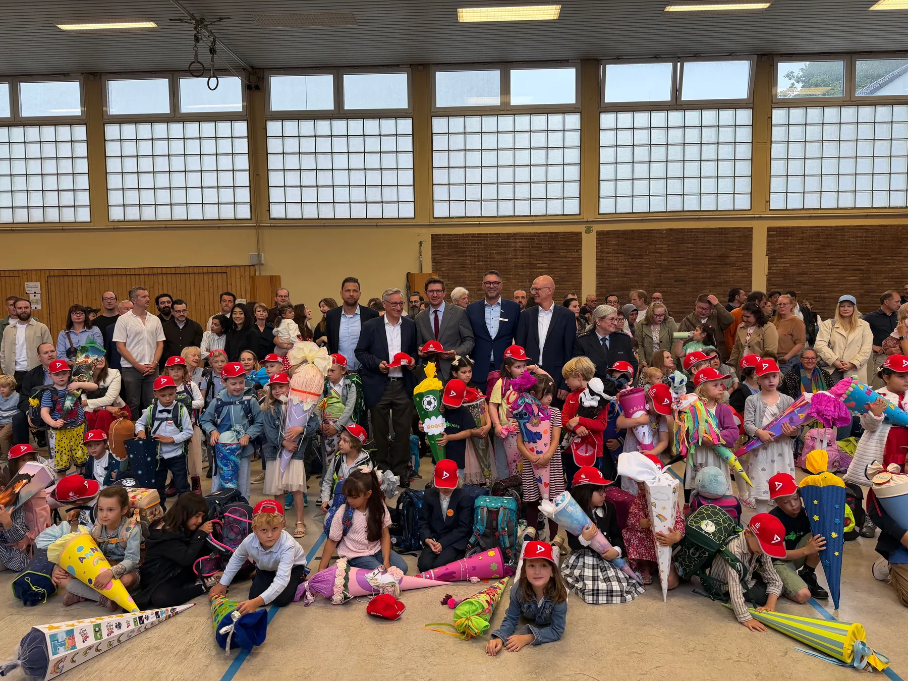 Viele Grundschulkinder mit bunten Schultüten und roten Kappen in Turnhalle, Erwachsene im Hintergrund.