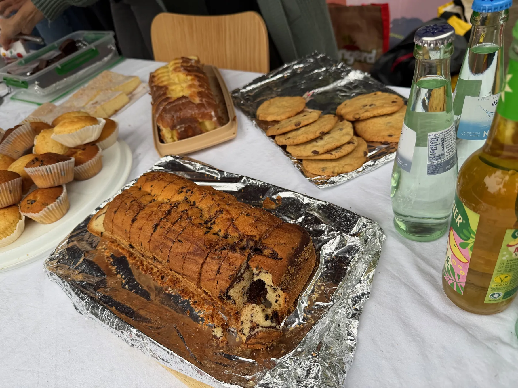 Angeschnittener Marmorkuchen auf Alufolie, daneben Muffins, Schokoladenkekse und Flaschen auf Tisch