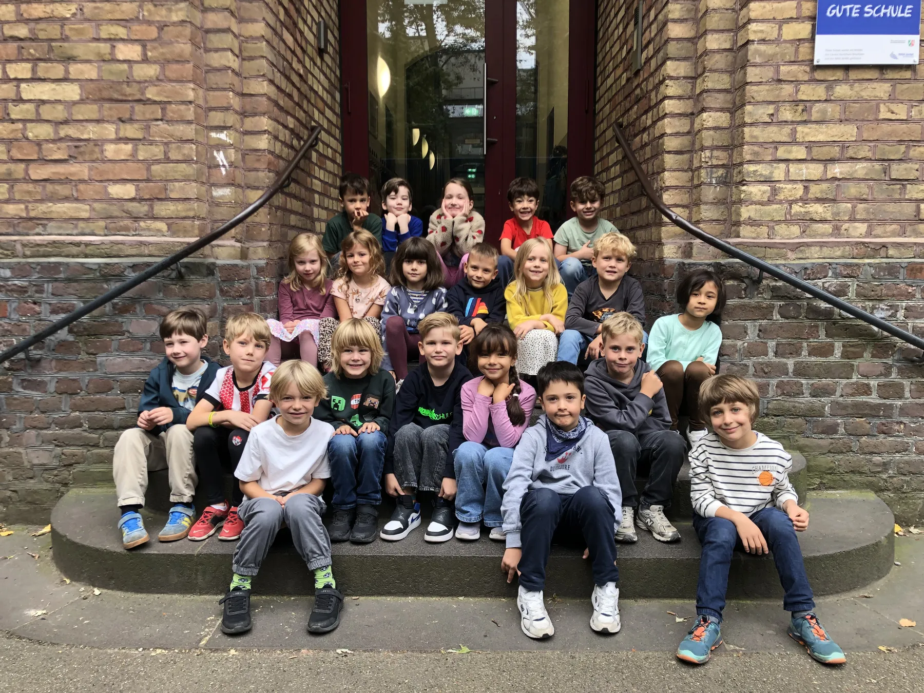Gruppe von Kindern sitzt auf Treppenstufen vor einem Backsteingebäude mit Glas-Eingang
