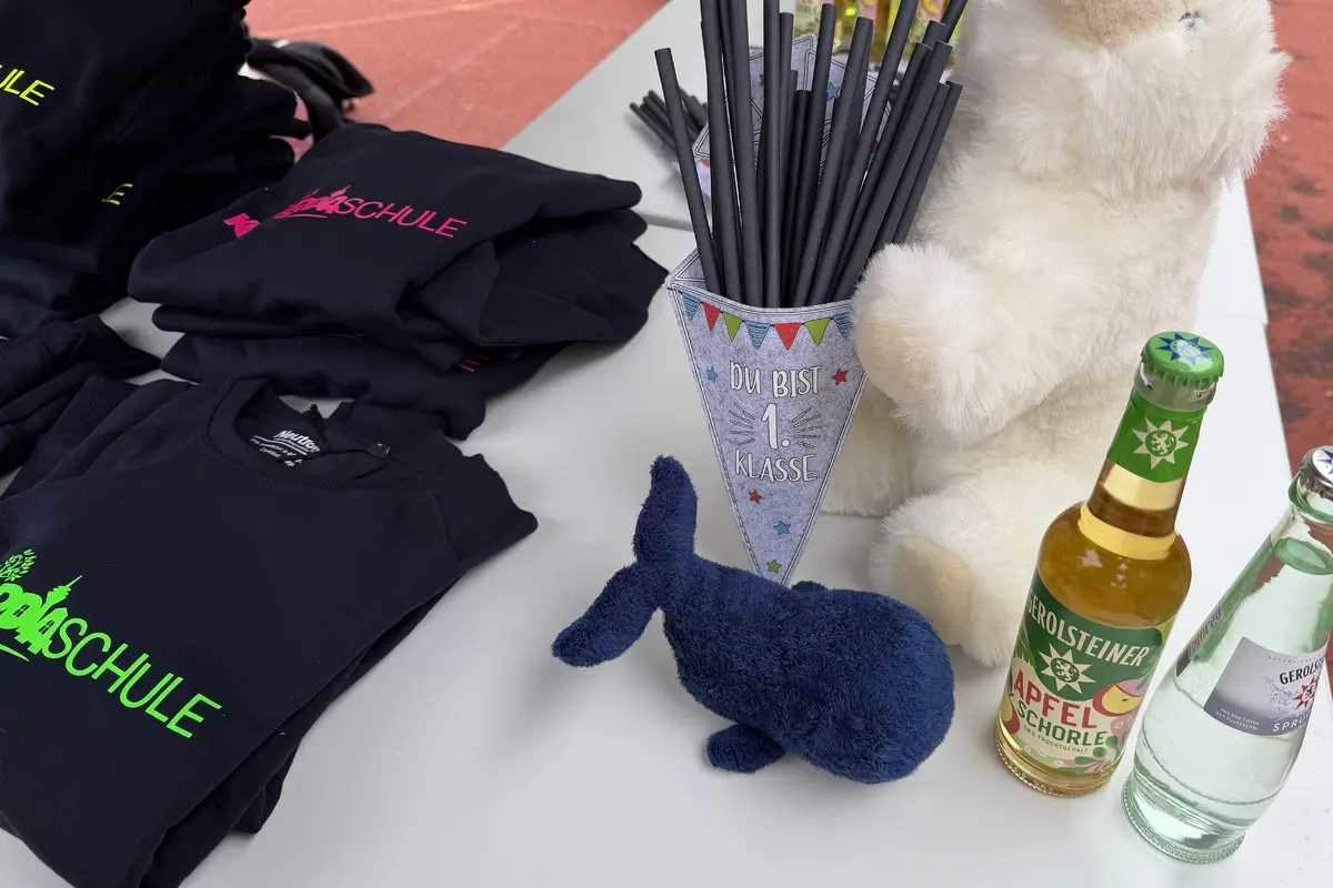 Gestapelte schwarze Schultshirts, Wimpel-"Du bist 1. Klasse" mit schwarzen Strohhalmen, weißer Plüschbär, blaues Stofftier und Getränkeflaschen