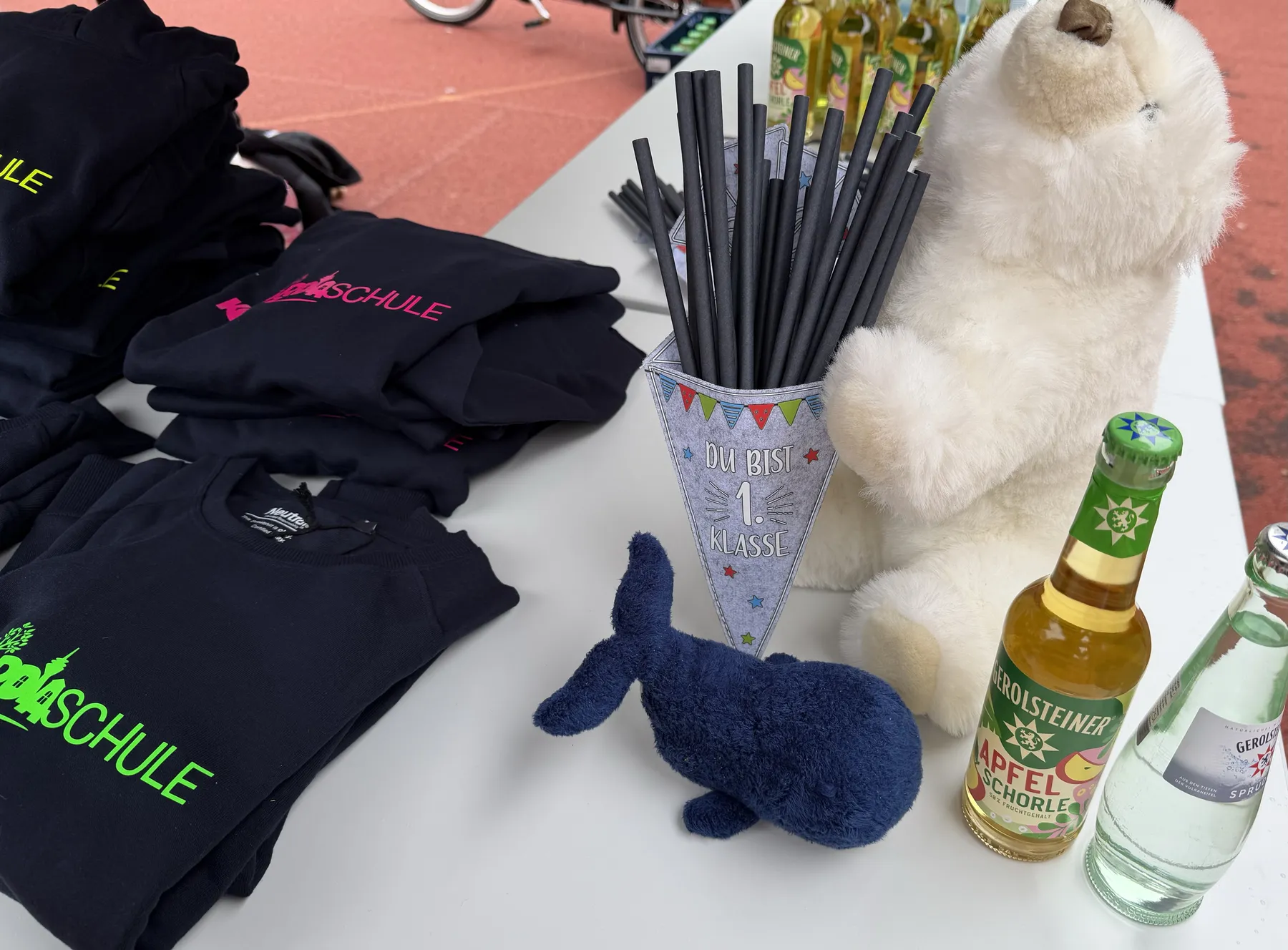 Gestapelte schwarze Schultshirts, Wimpel-"Du bist 1. Klasse" mit schwarzen Strohhalmen, weißer Plüschbär, blaues Stofftier und Getränkeflaschen