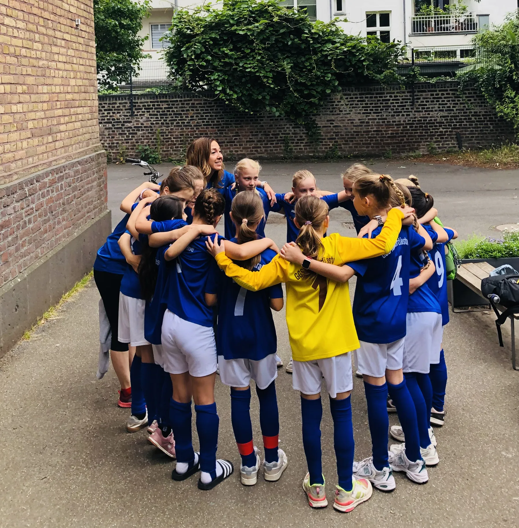Mädchen-Fußballteam in blauen Trikots umarmt sich im Kreis mit Trainerin, eine Spielerin in gelbem Trikot