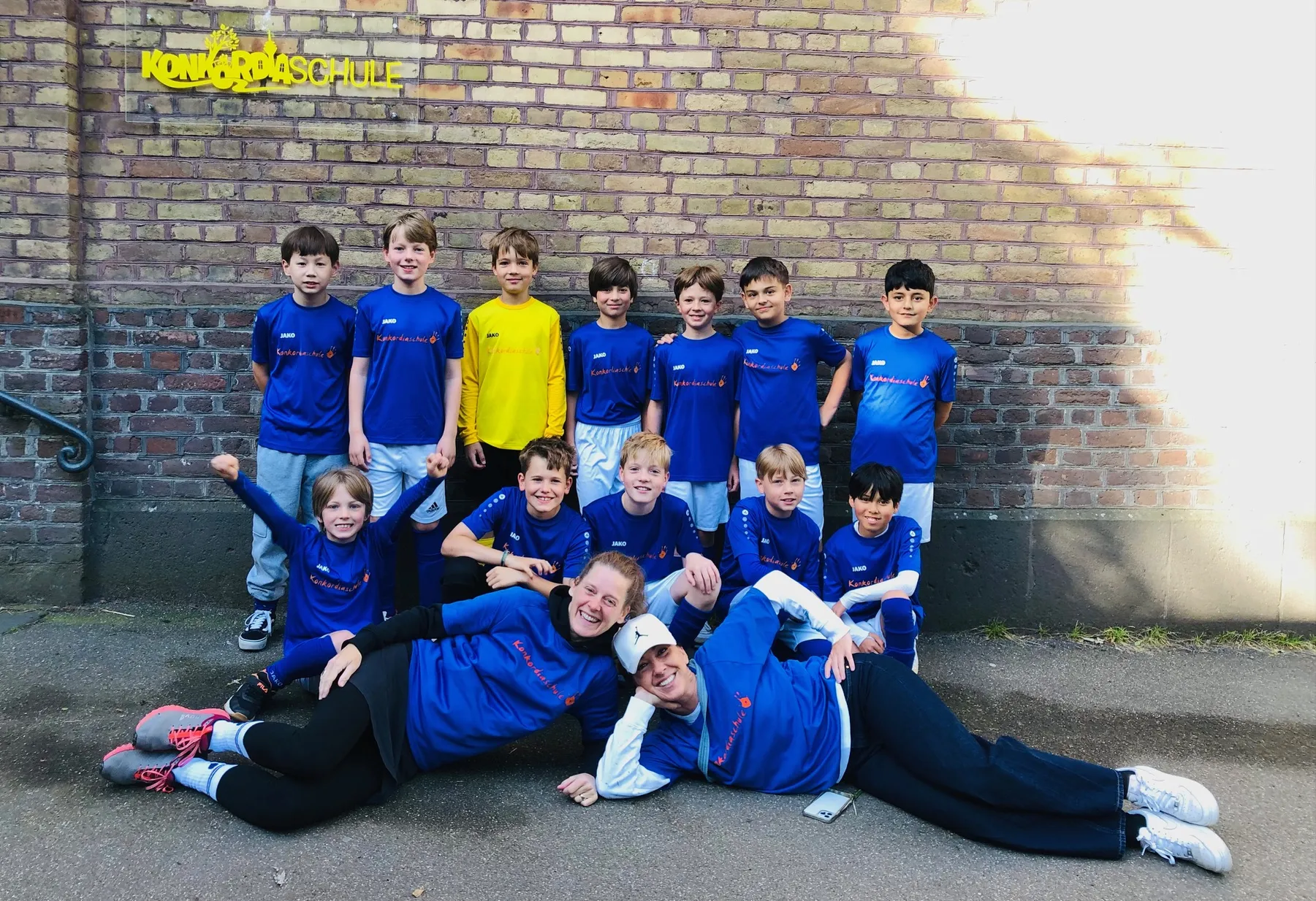 Kinder- und Erwachsenenteam in blauen Fußballtrikots posiert vor Backsteinwand mit gelbem "Konkordiaschule"-Schriftzug.