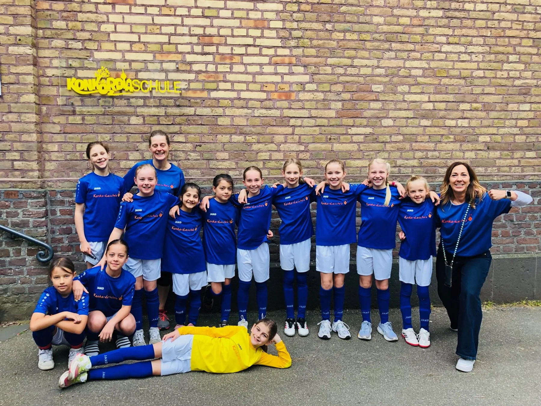 Elf Mädchen in blauen Fußballtrikots mit Trainerin vor Backsteinwand, eine Spielerin liegt vorn in gelbem Trikot