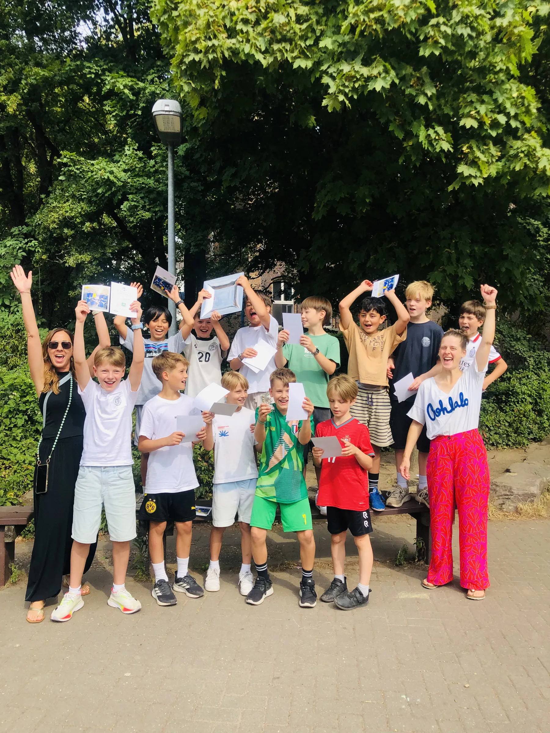 Fröhliche Gruppe Kinder und zwei Erwachsene halten Urkunden und einen Pokal hoch und jubeln vor großen Bäumen