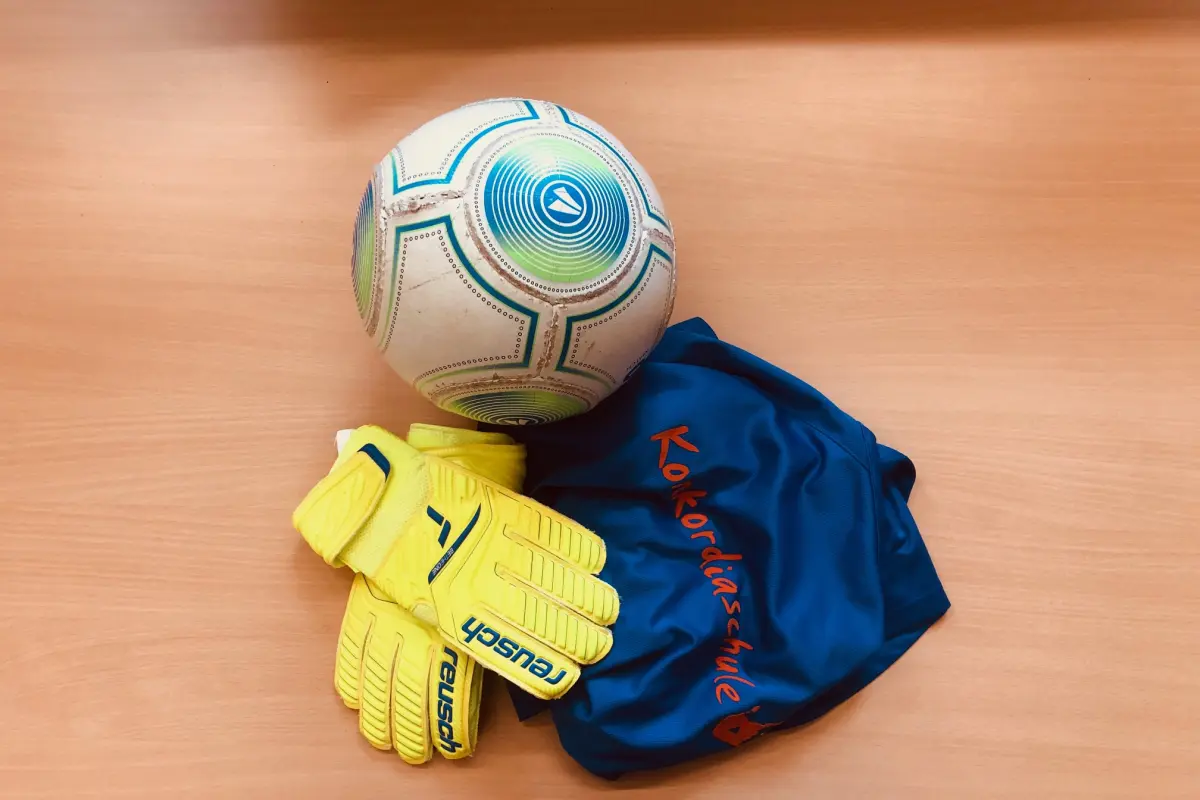 Weiß-blauer Fußball, gelbe Torwarthandschuhe und blaues Trikot mit orangefarbener Schrift auf hellem Holztisch