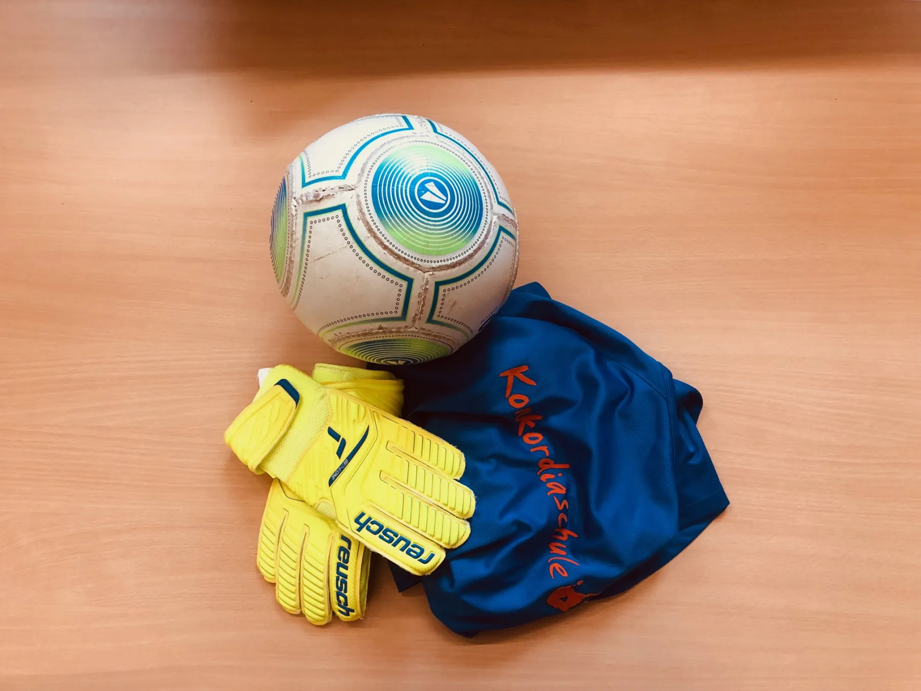Weiß-blauer Fußball, gelbe Torwarthandschuhe und blaues Trikot mit orangefarbener Schrift auf hellem Holztisch