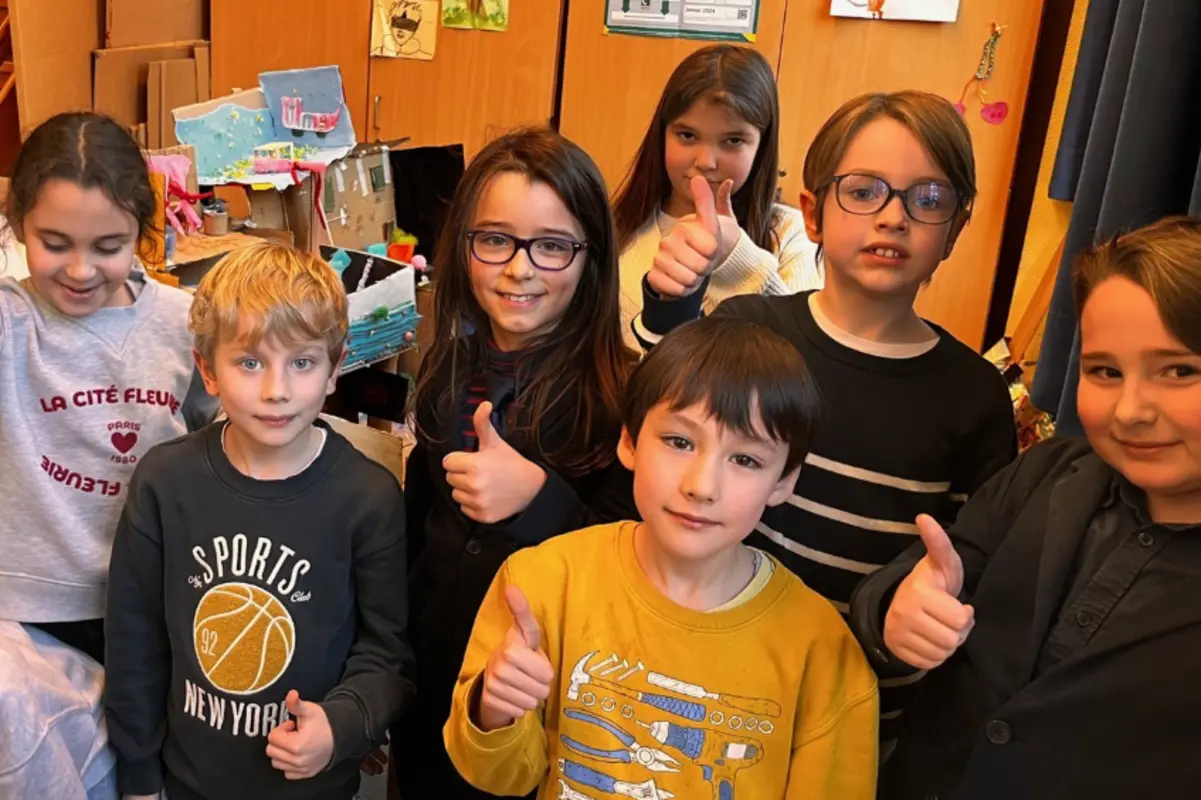Sieben Kinder in einem Klassenraum zeigen Daumen hoch und lächeln.