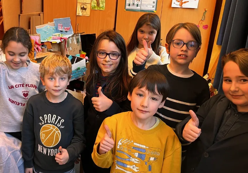 Sieben Kinder in einem Klassenraum zeigen Daumen hoch und lächeln.