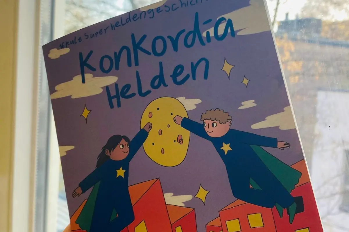 Hand hält Comic mit Titel „Konkordia Helden“, zwei fliegende, sternbekleidete Figuren über roten Hochhäusern vor Fensterkulisse