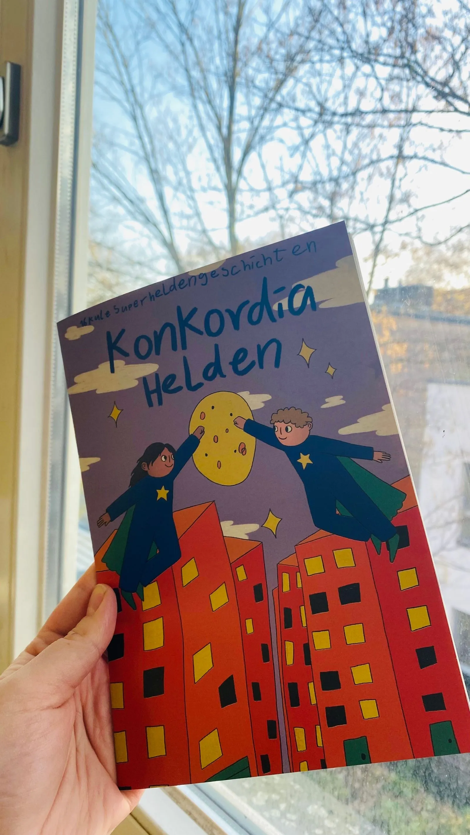 Hand hält Comic mit Titel „Konkordia Helden“, zwei fliegende, sternbekleidete Figuren über roten Hochhäusern vor Fensterkulisse