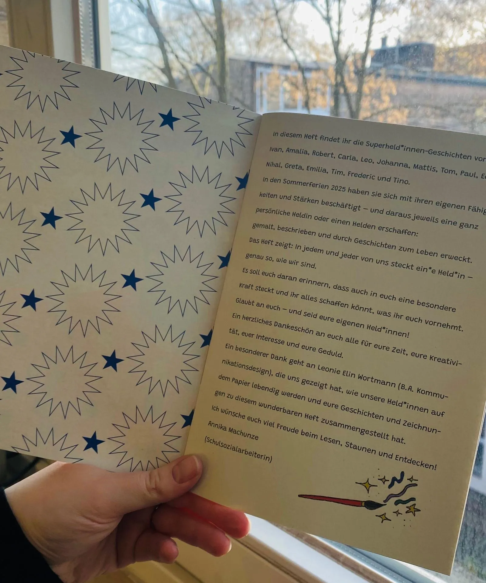 Hand hält aufgeklapptes Heft vor Fenster; linke Seite Muster mit blauen Sternen, rechte Seite Text und Pinselillustration.