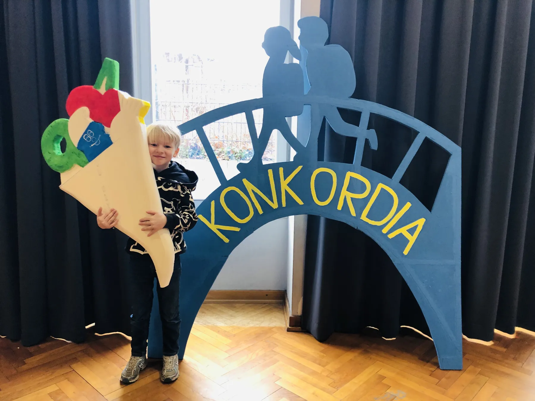 Kind hält große bunte Schultüte neben blauem Dekobogen mit gelber Aufschrift "KONKORDIA"