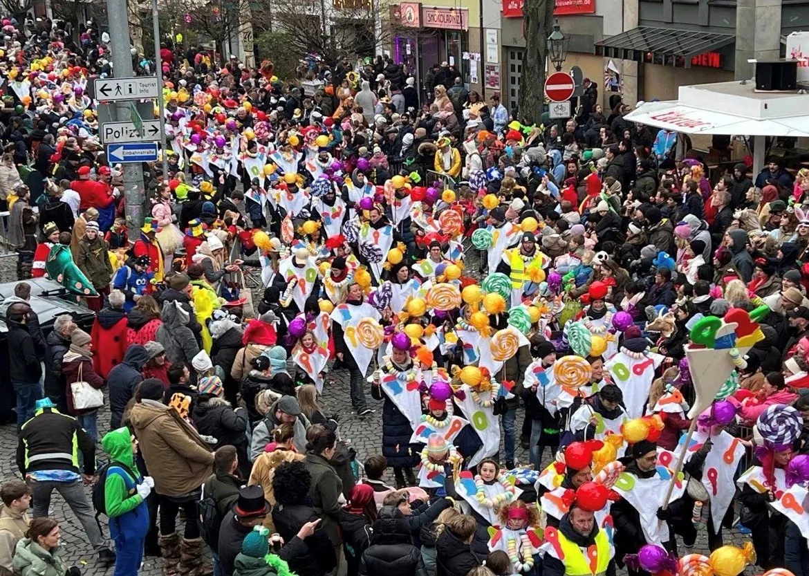 Bunter Karnevalsumzug: viele verkleidete Menschen mit übergroßen Lutschern und bunten Ballons in einer Straße