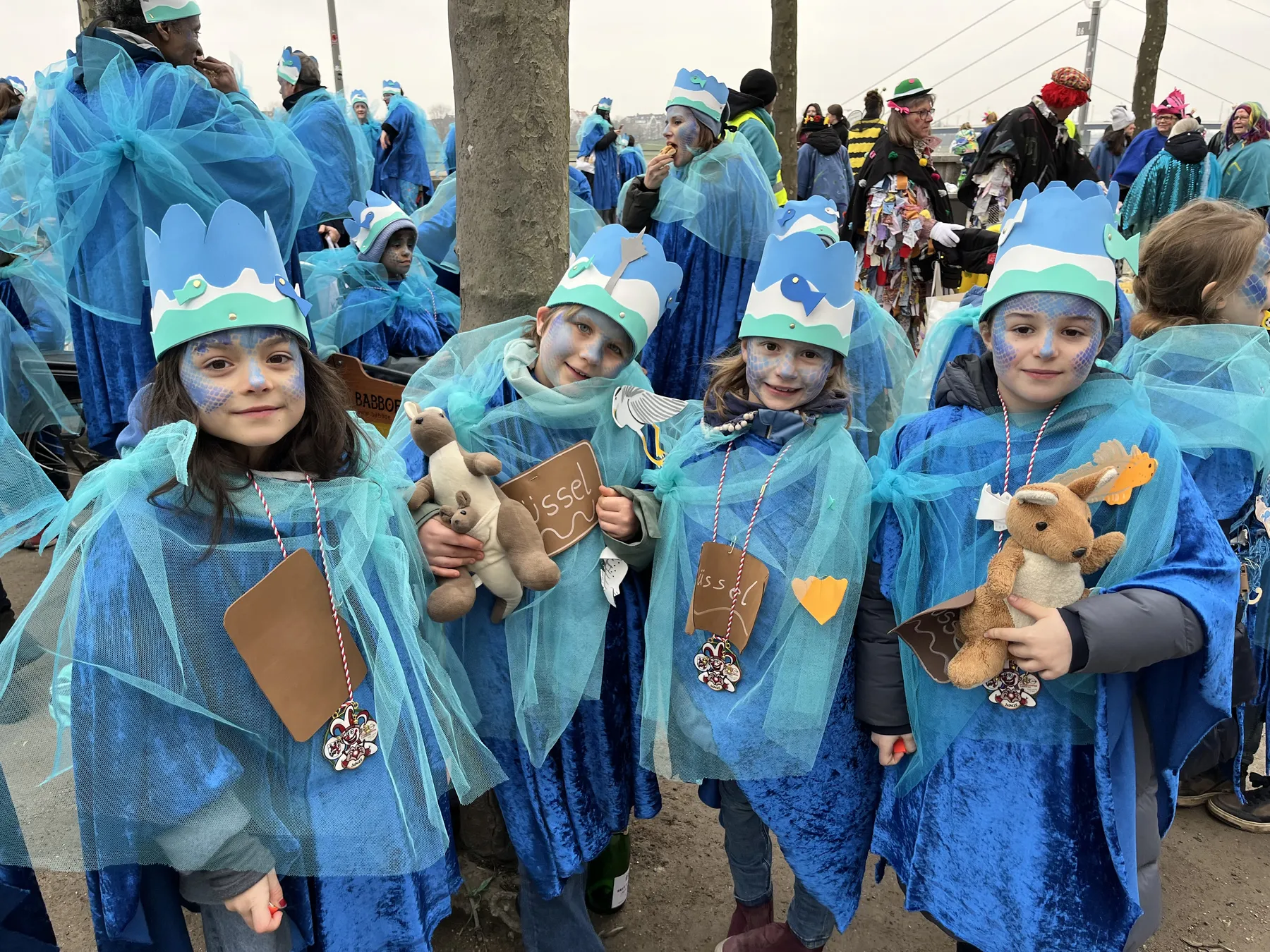 Kinder in blauen Meerjungfrauen‑Kostümen mit Papierkronen, blauem Netzstoff, Gesichtsfarbe und Kuscheltieren