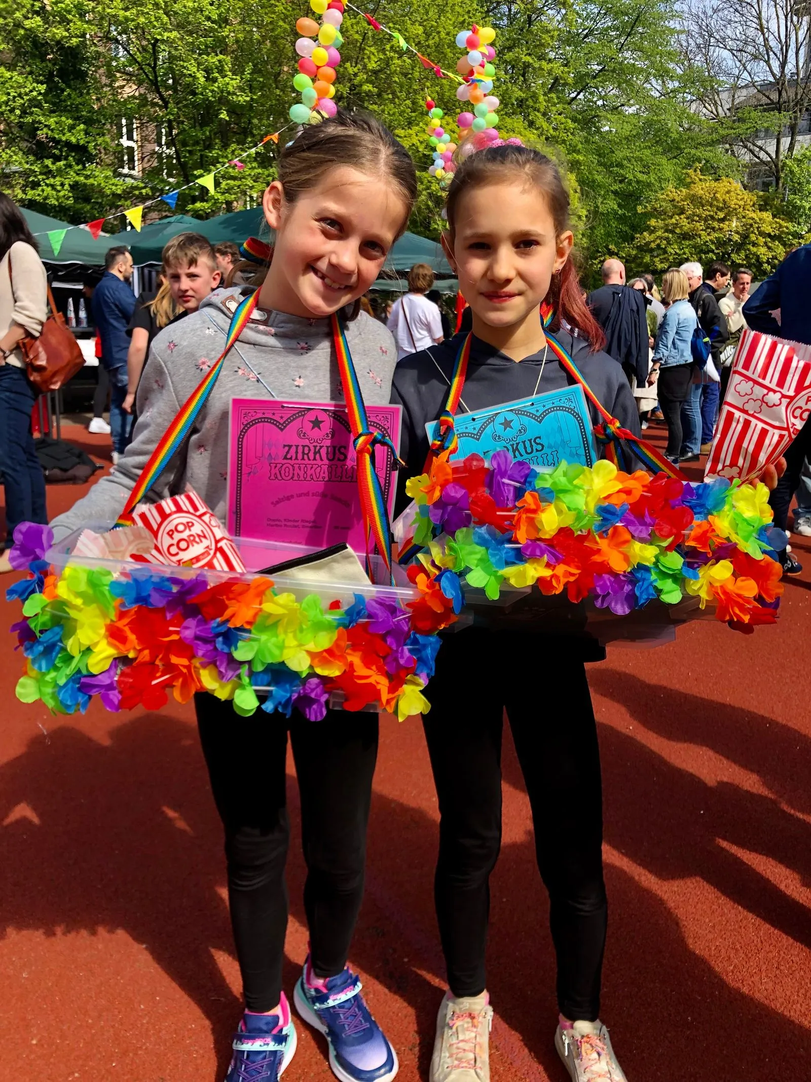 Zwei Mädchen mit Regenbogenbändern halten bunte Blumengirlanden, Popcorn und Zirkus-Zertifikate auf einem Fest
