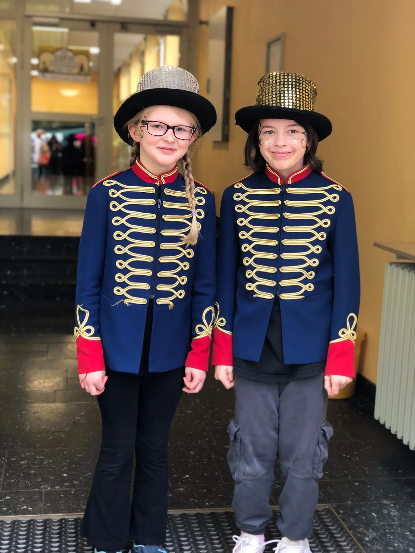 Zwei Kinder in blauen Paradejacken mit goldener Borte und glitzernden Zylindern, lächelnd mit Gesichtsbemalung