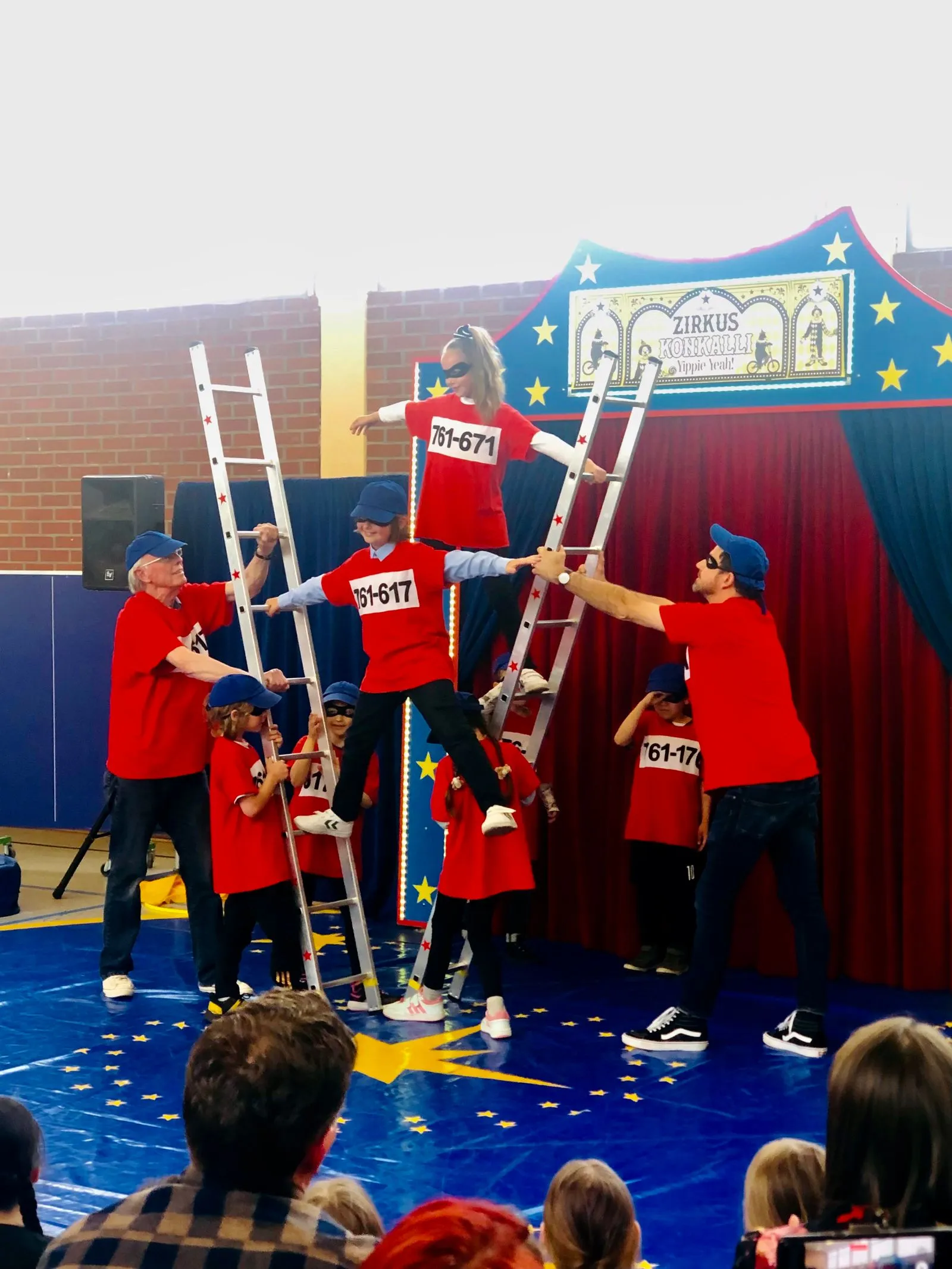 Kinder und Erwachsene in roten Shirts bauen eine Leiterpyramide vor blau-roter Zirkuskulisse