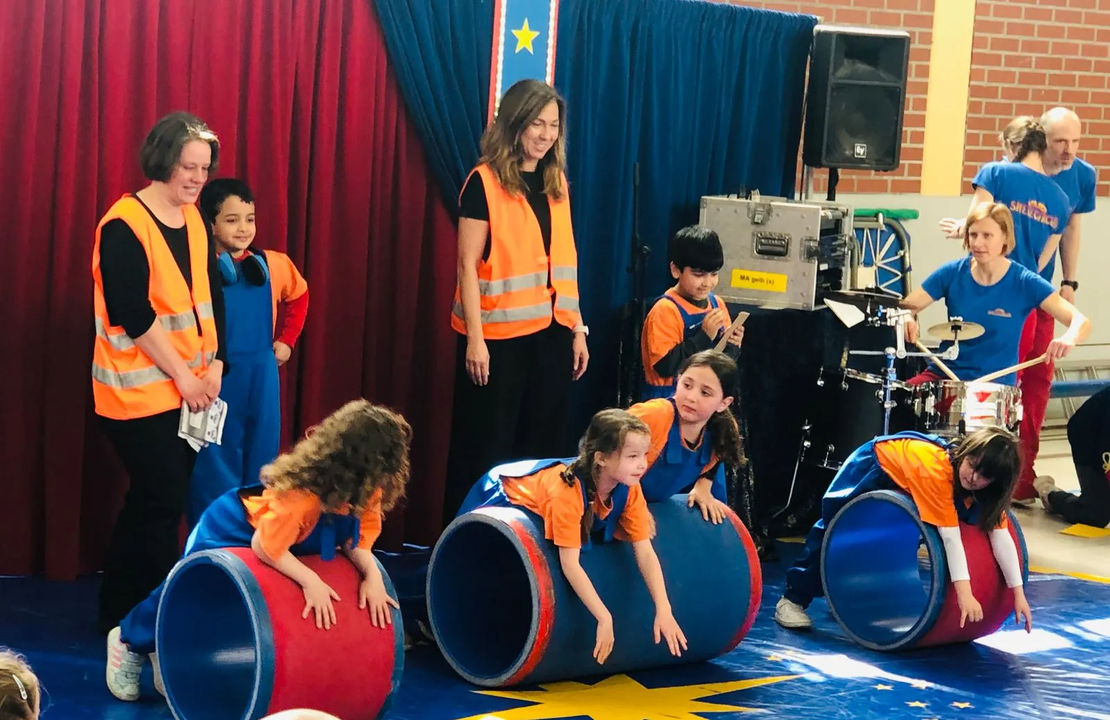 Kinder in blauen Overalls und orangen T-Shirts rollen auf großen roten/blauen Zylindern, Betreuer in Warnwesten daneben.