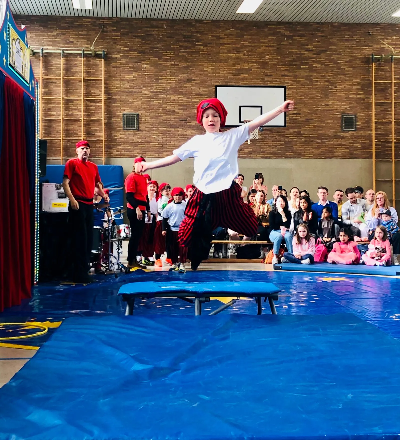 Kind mit rotem Kopftuch und weißem T‑Shirt springt über Trampolin vor zuschauender Menschenmenge in Sporthalle