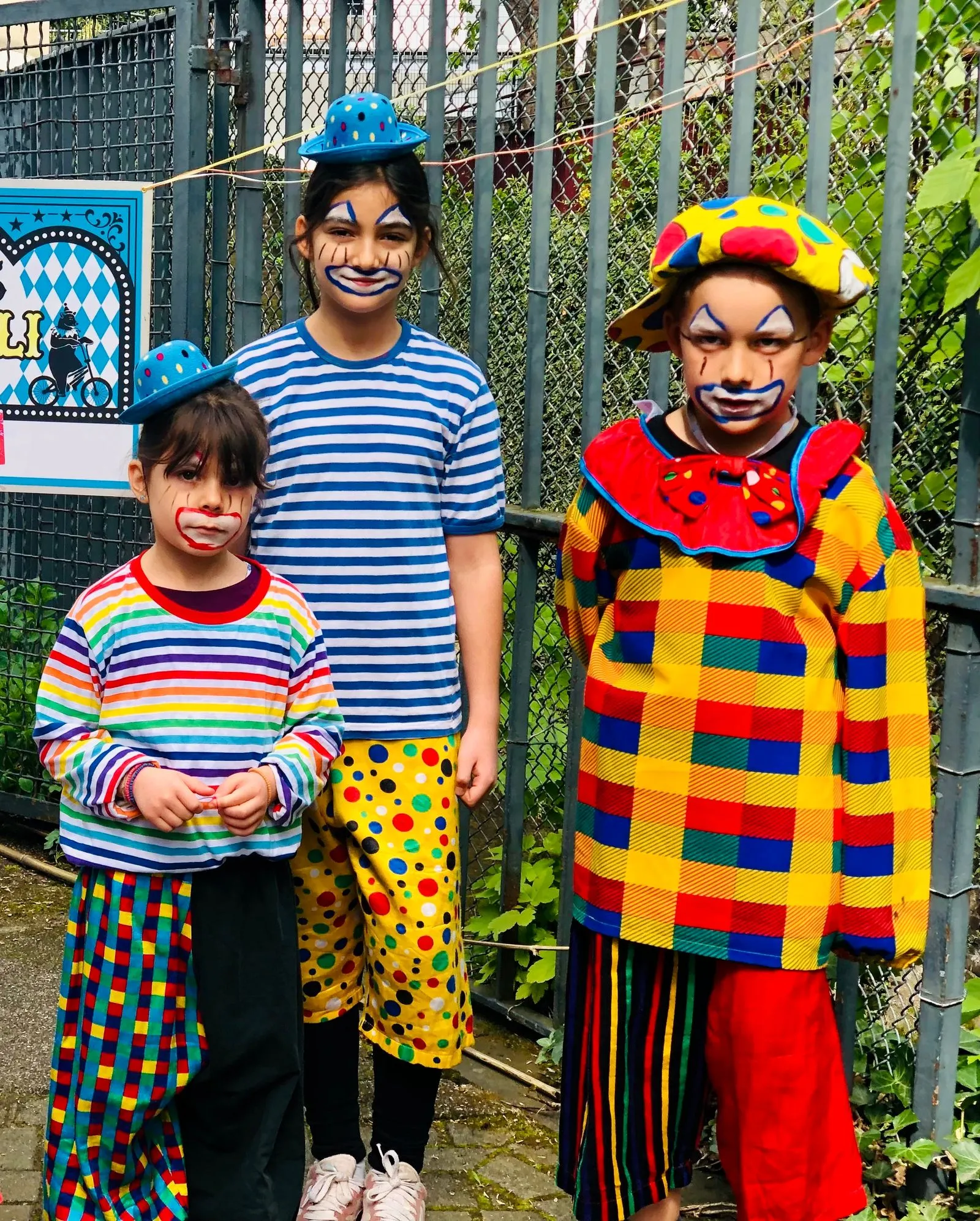 Drei verkleidete Kinder mit Clownsmakeup in bunten gestreiften und karierten Kostümen vor Maschendrahtzaun