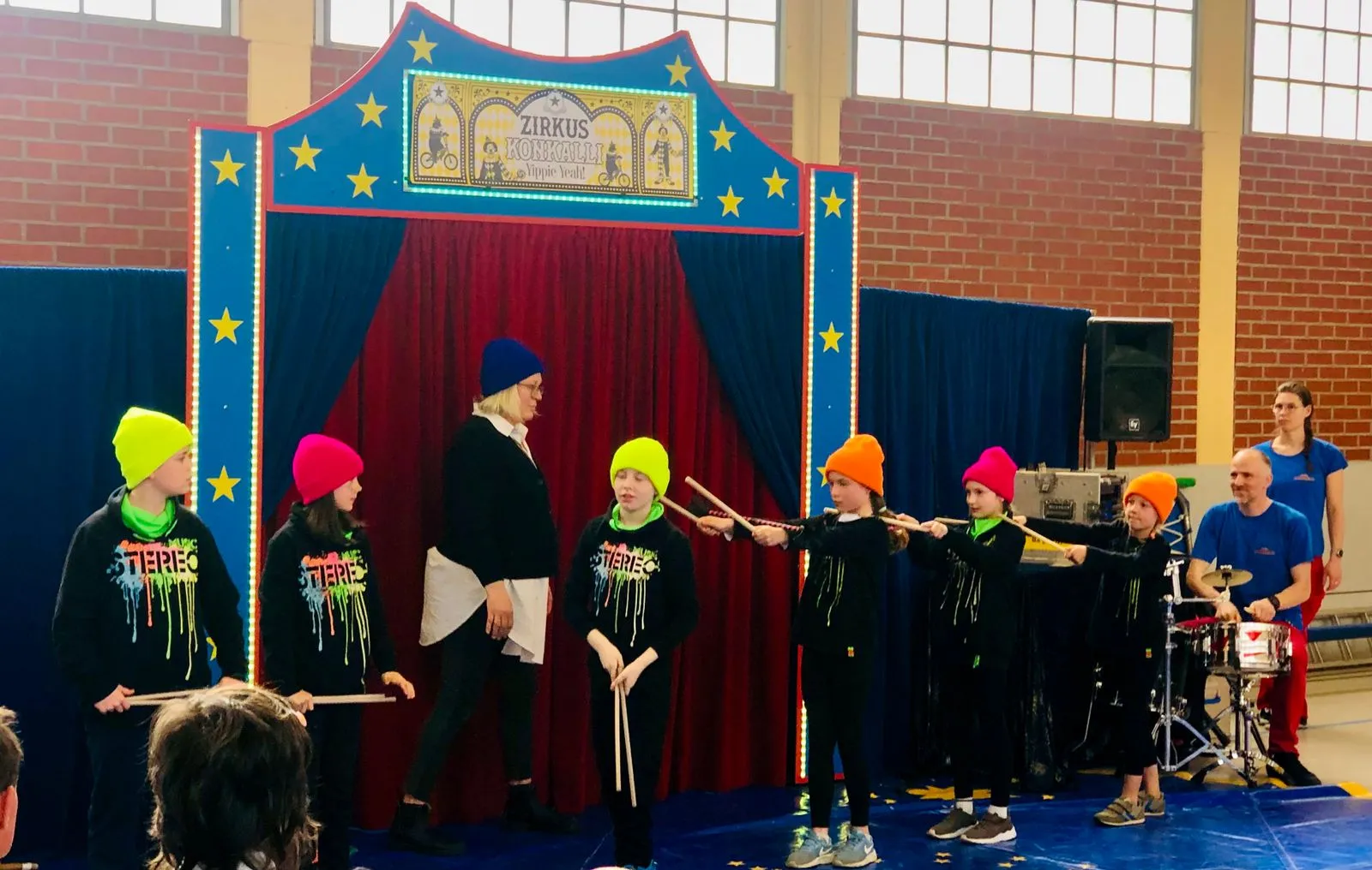 Kinder in neonfarbigen Mützen und schwarzen Shirts halten Schlagstöcke vor blau-roter Zirkusbühne, rechts Erwachsener am Schlagzeug