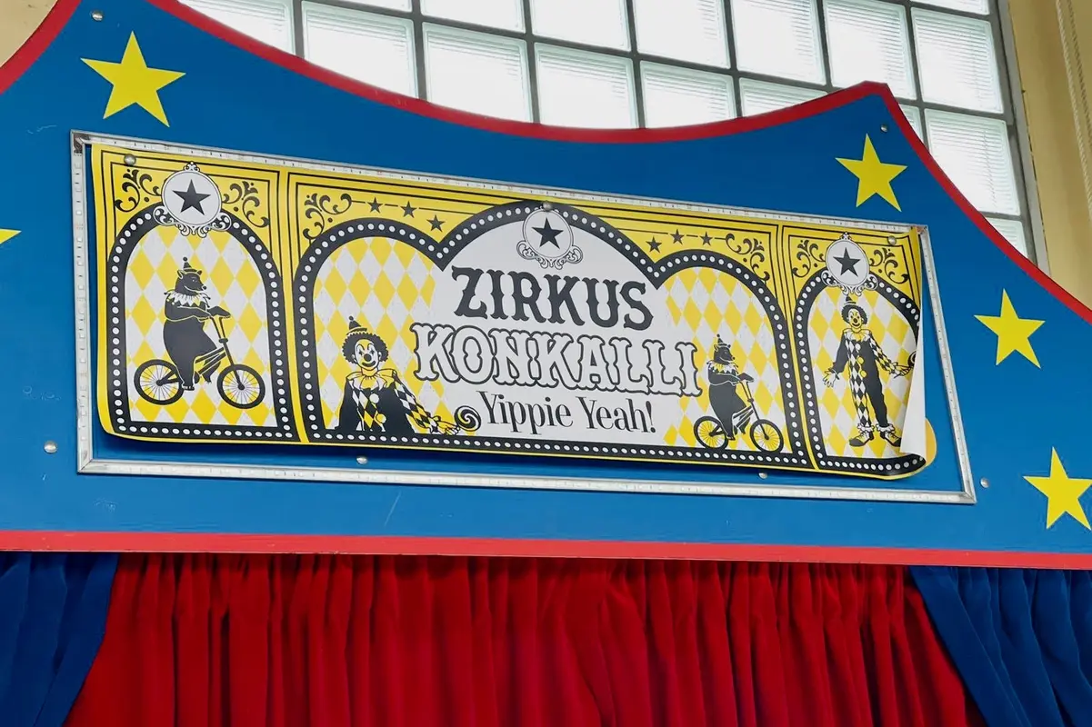 Zirkusschild mit Schrift "ZIRKUS KONIKAILLI", Clowns und Bären auf Fahrrädern über rotem Vorhang