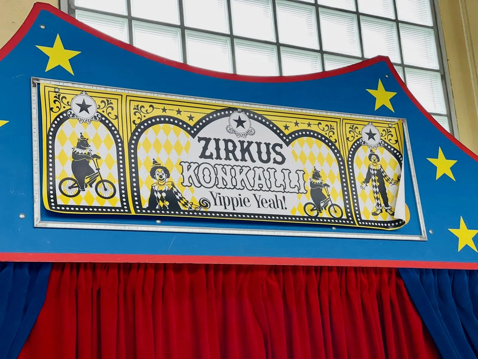 Zirkusschild mit Schrift "ZIRKUS KONIKAILLI", Clowns und Bären auf Fahrrädern über rotem Vorhang