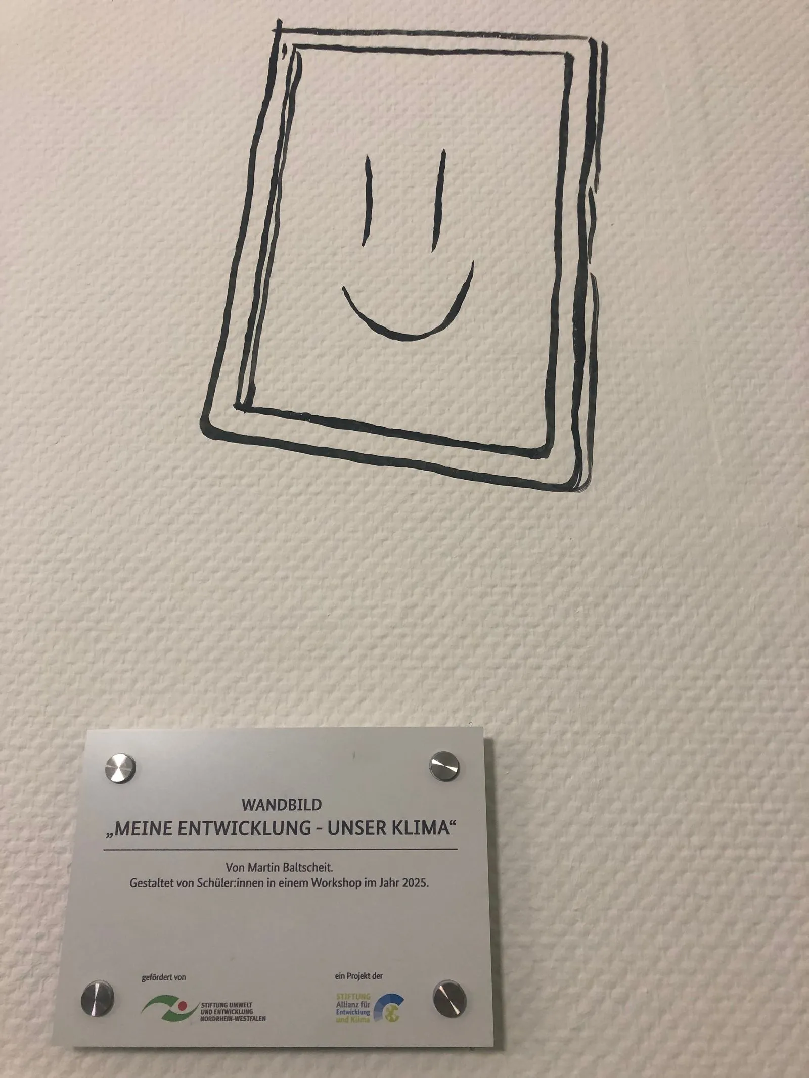 Schwarz gezeichnetes, umrahmtes Smiley über beschrifteter Metalltafel an strukturierter weißer Wand