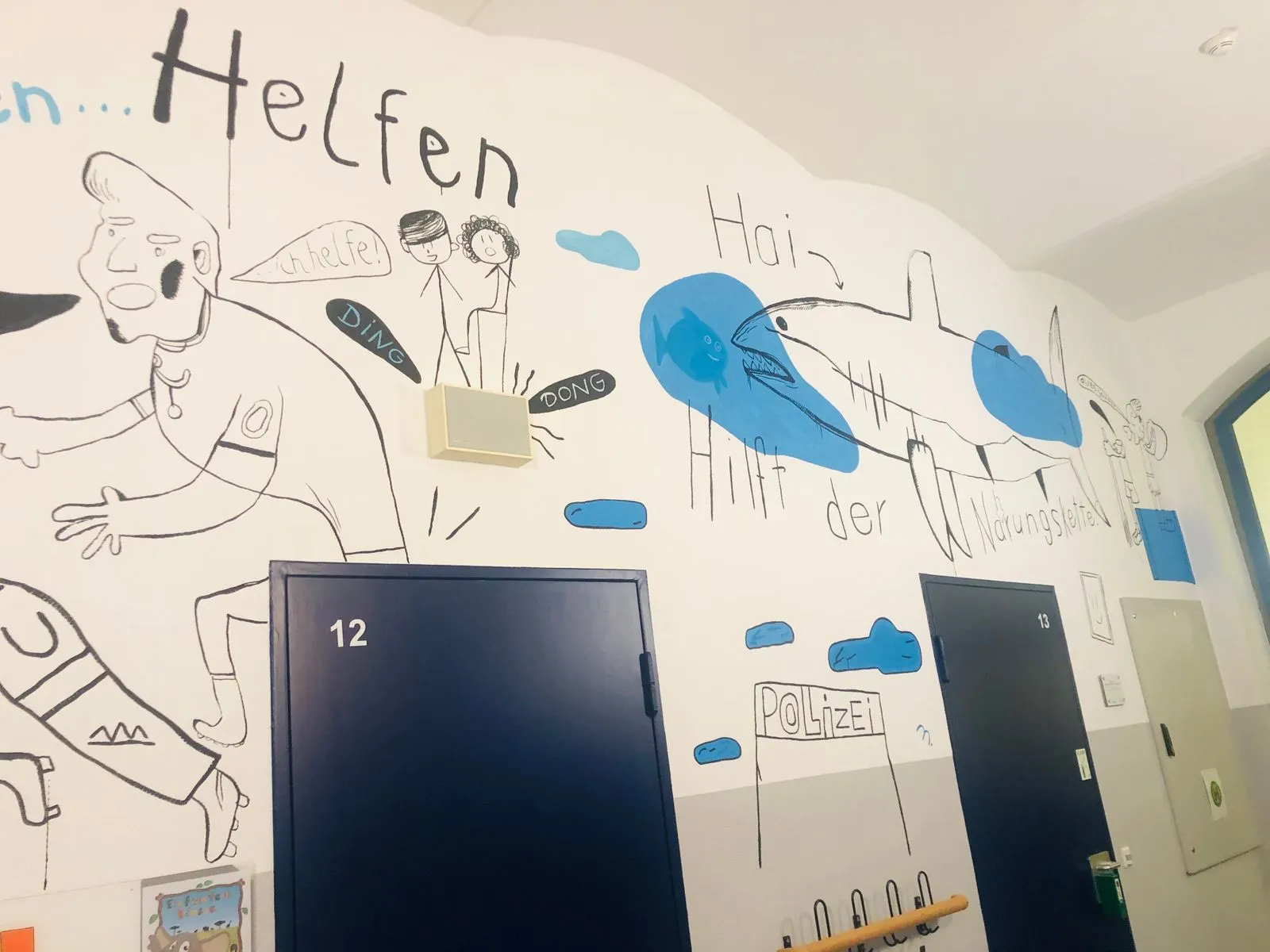 Weiße Wand mit schwarzen Comic-Zeichnungen, blau akzentuierten Formen und Hai sowie zwei dunkelblaue Türen mit Nummern 12 und 13