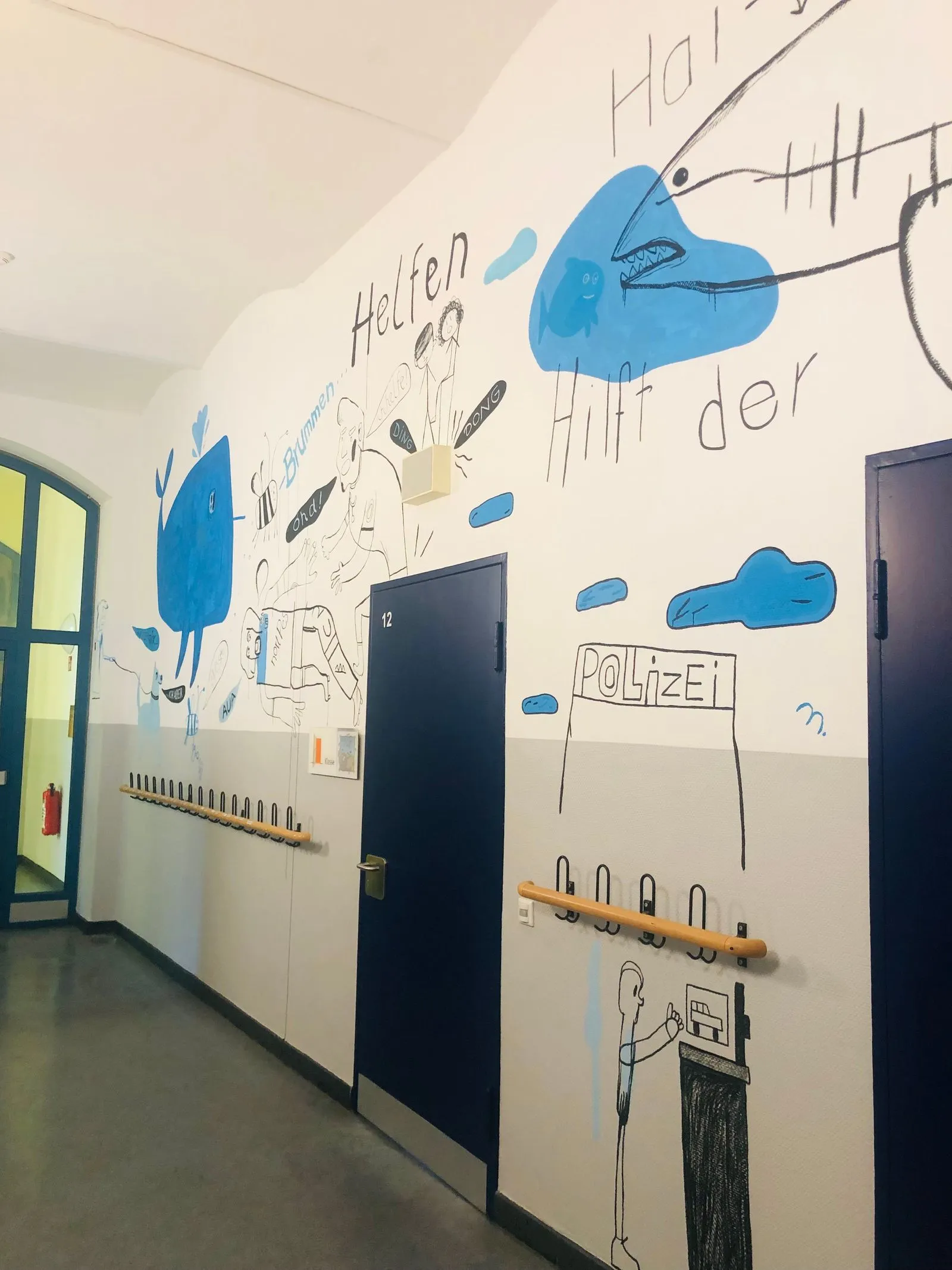 Flur mit dunkelblauen Türen, Handläufen und Wandbemalung mit blauen Wolken, Figuren und der Aufschrift "Polizei"