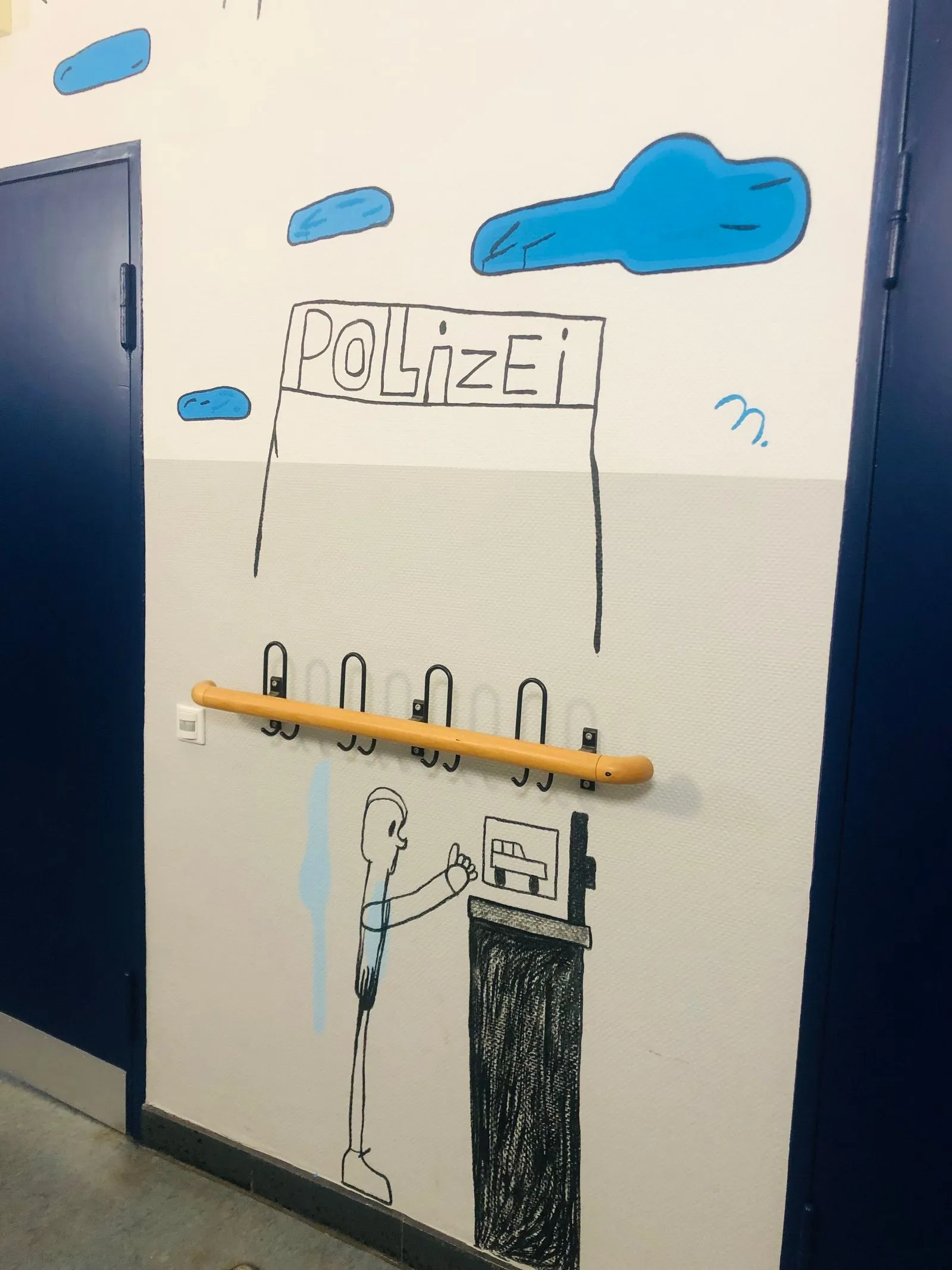 Wandzeichnung mit "POLIZEI"-Schild, blauen Wolken, gelbem Handlauf, Garderobenhaken und gezeichnetem Menschen am Automaten.