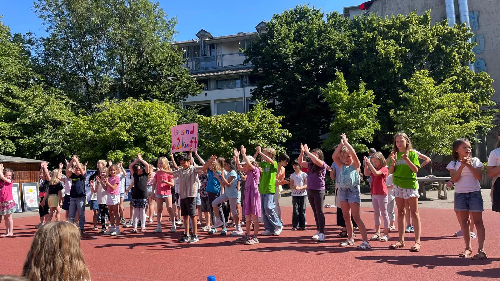 Kinder auf Schulhof klatschen, eines hält pinkes Schild „Wir sind Zukunft“, Bäume und Wohngebäude im Hintergrund