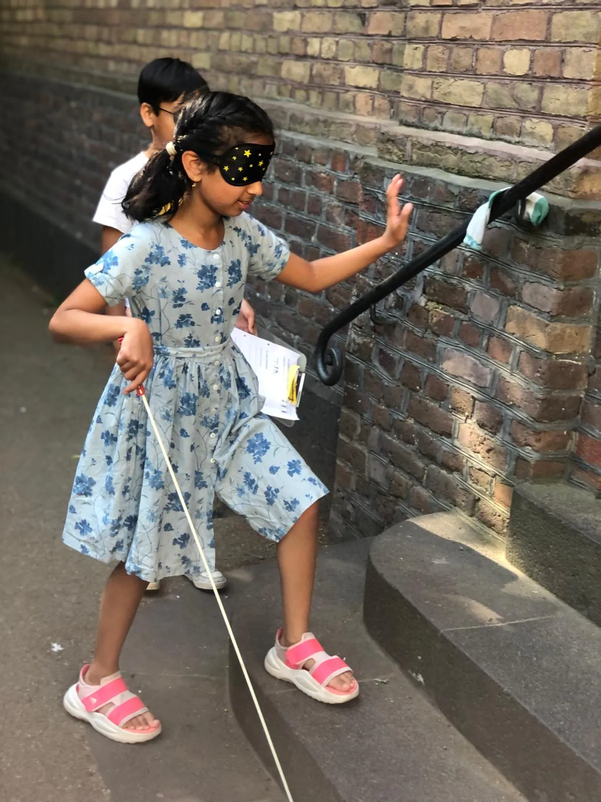 Mädchen mit sternenbedruckter Augenbinde und weißem Langstock tastet Hand an Treppengeländer, trägt blaues Blumenkleid
