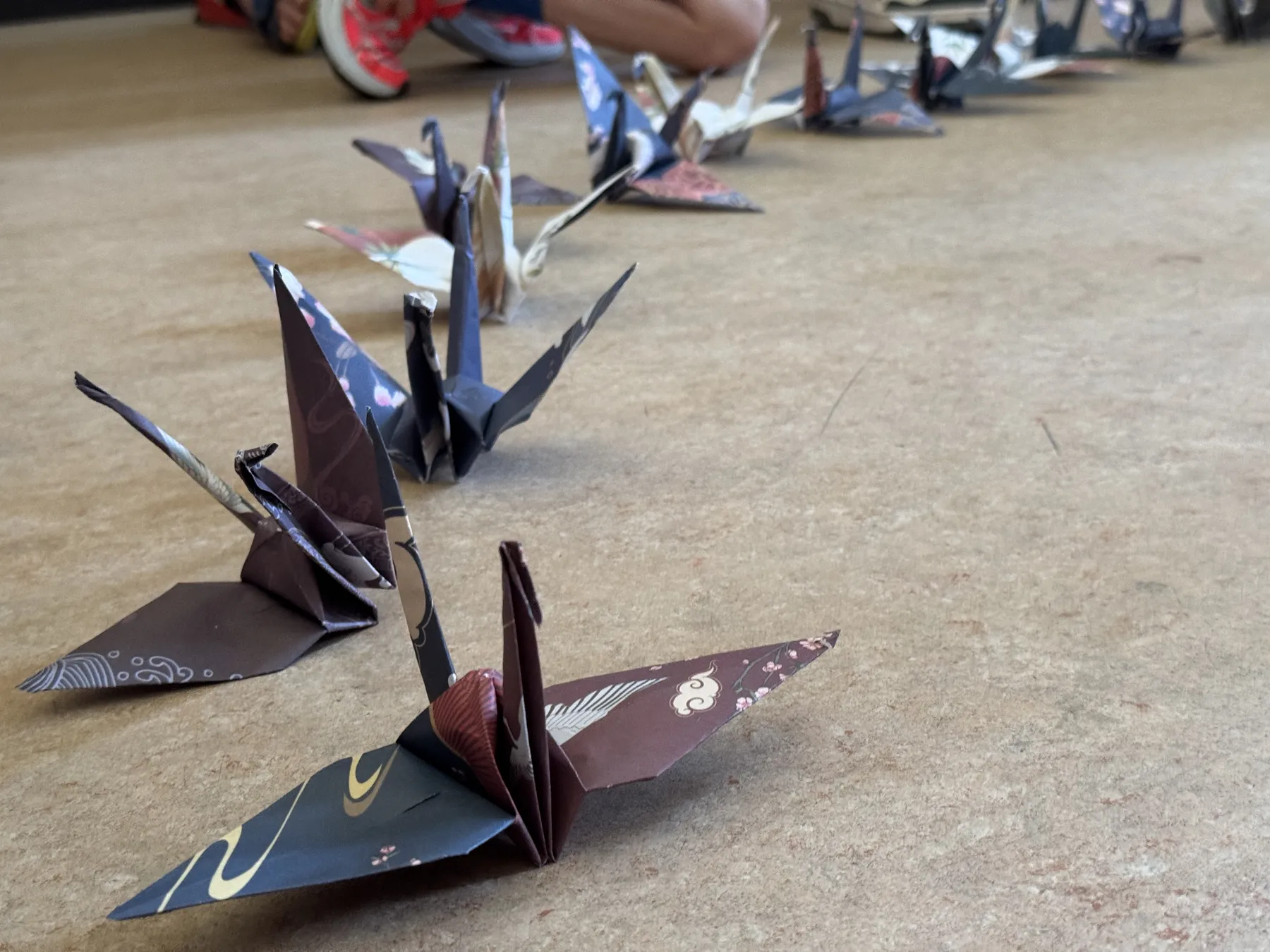 Mehrere bunte Origami-Kraniche in Reihe auf dem Boden, unscharfe Beine und rote Turnschuhe im Hintergrund