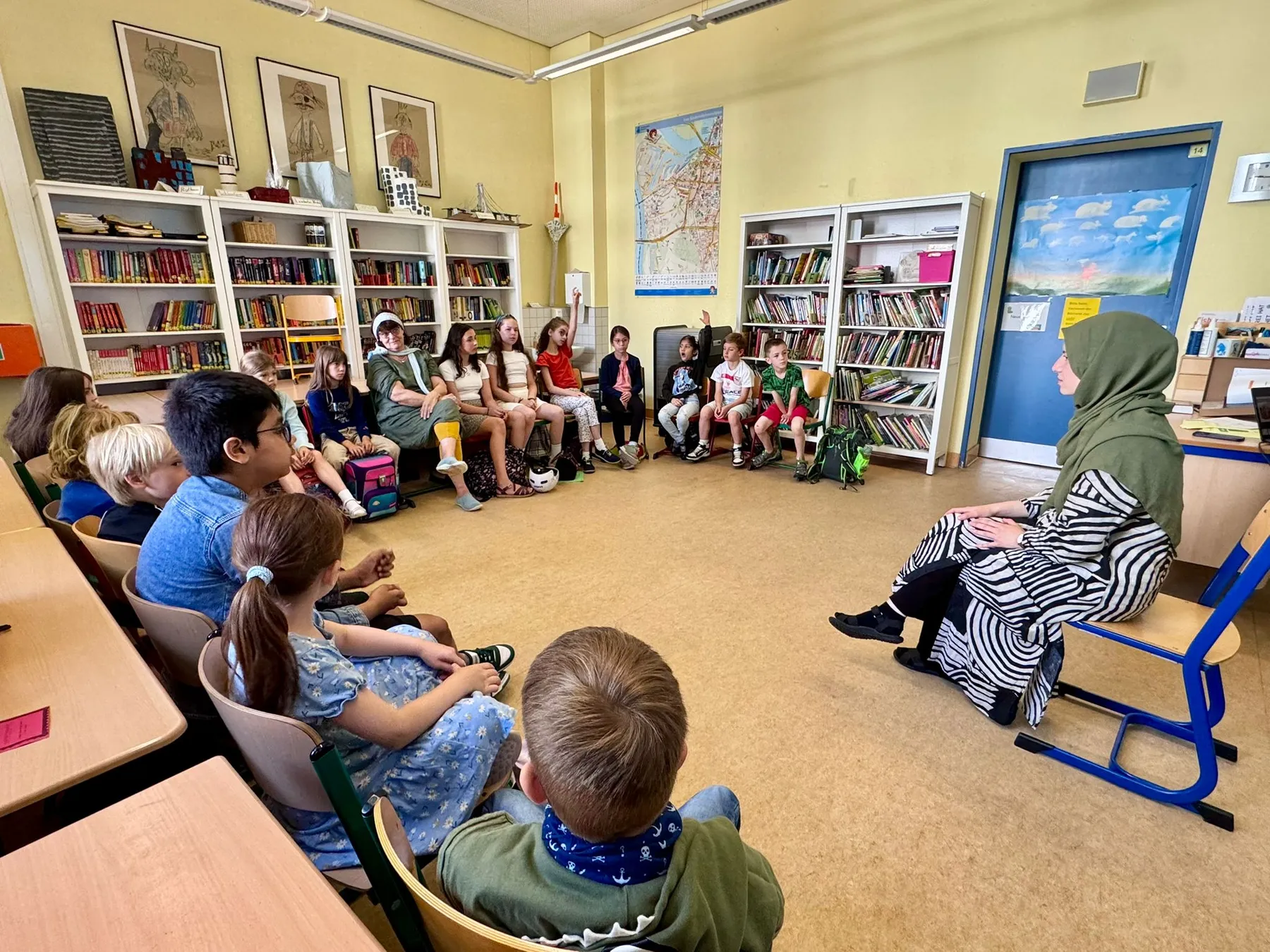 Grundschüler sitzen im Stuhlkreis in einer Bibliotheksklasse, Frau mit grünem Kopftuch spricht