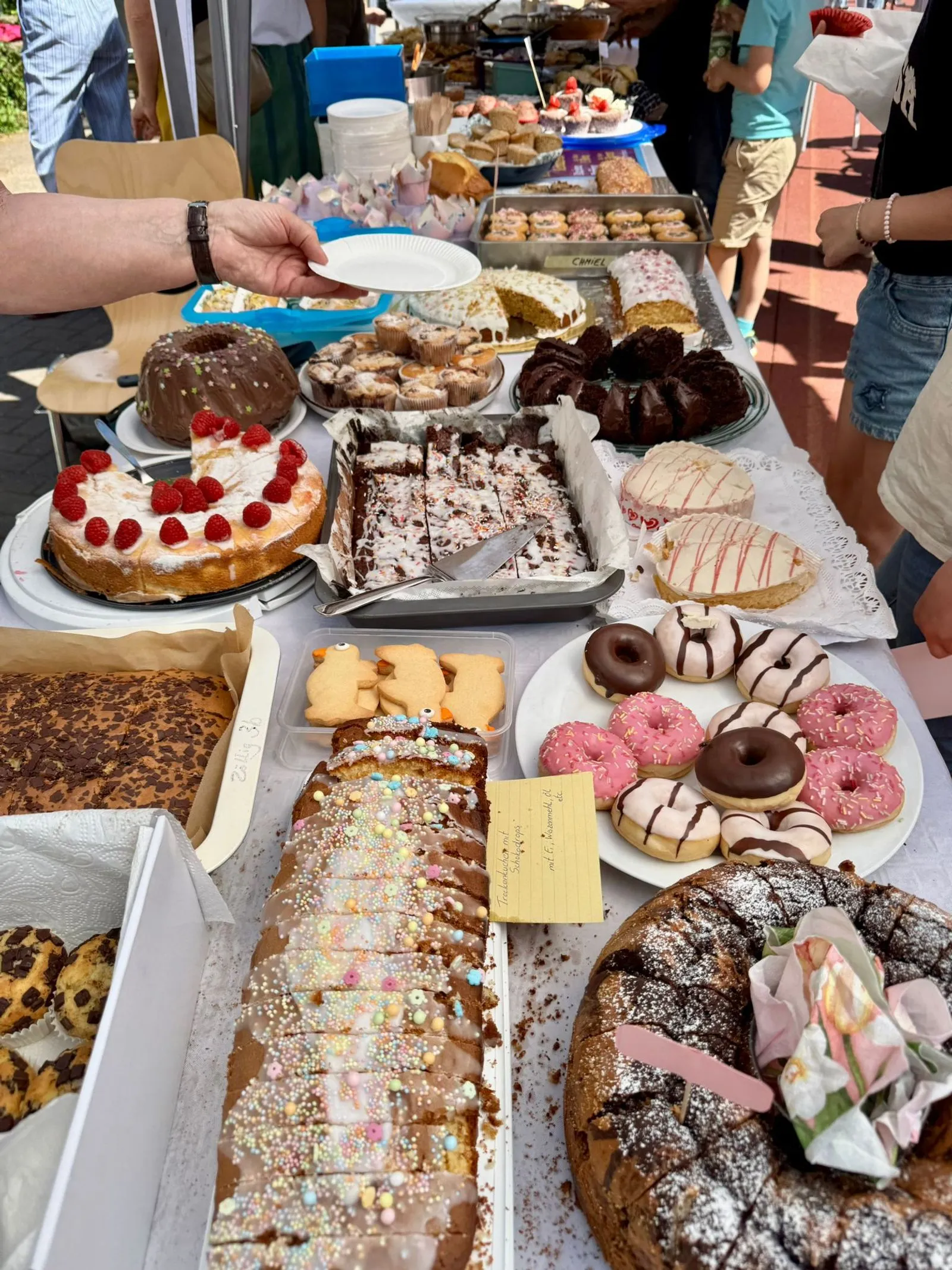 Tisch mit verschiedenen Kuchen, Donuts und Gebäck; eine Hand hält einen leeren Papp­teller.