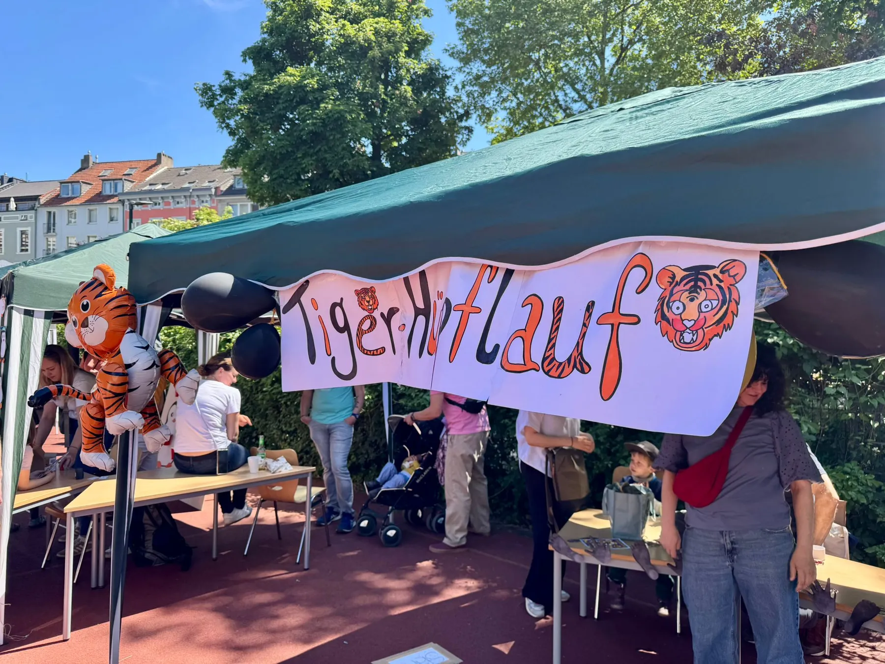 Grüner Pavillion mit Banner „Tigerhilfe“, Tiger-Luftballons und mehreren Personen darunter
